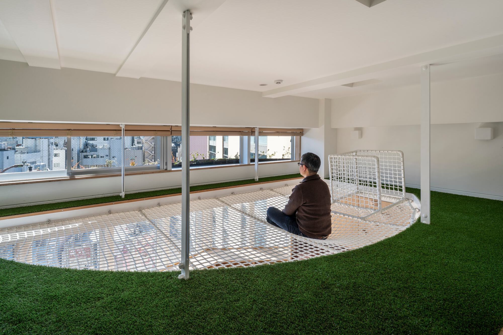 Person seated on the curved rope net floor beside a band of windows, surrounded by green turf