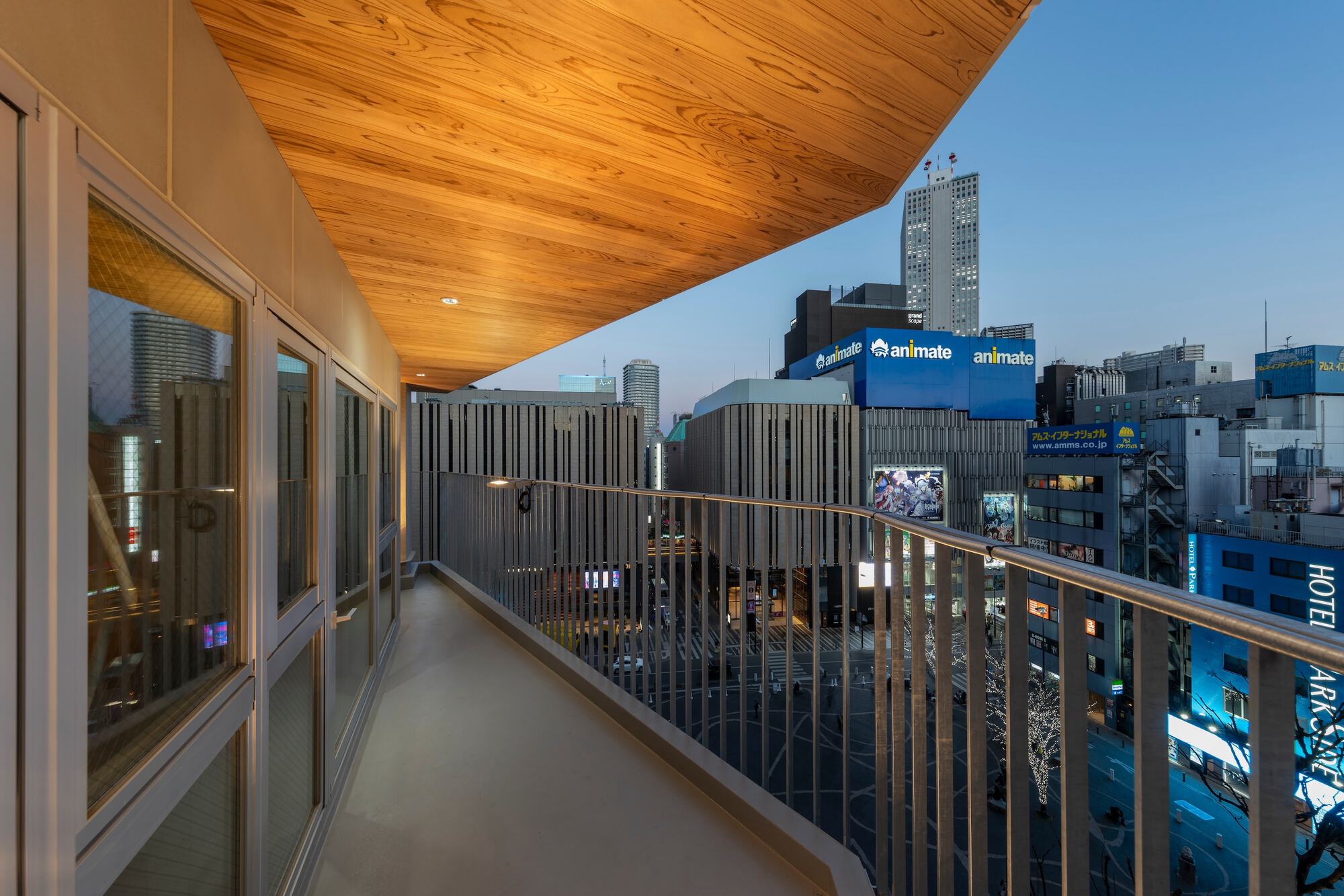 Narrow balcony with slatted railing and timber soffit overlooking a lit city intersection and signs at dusk