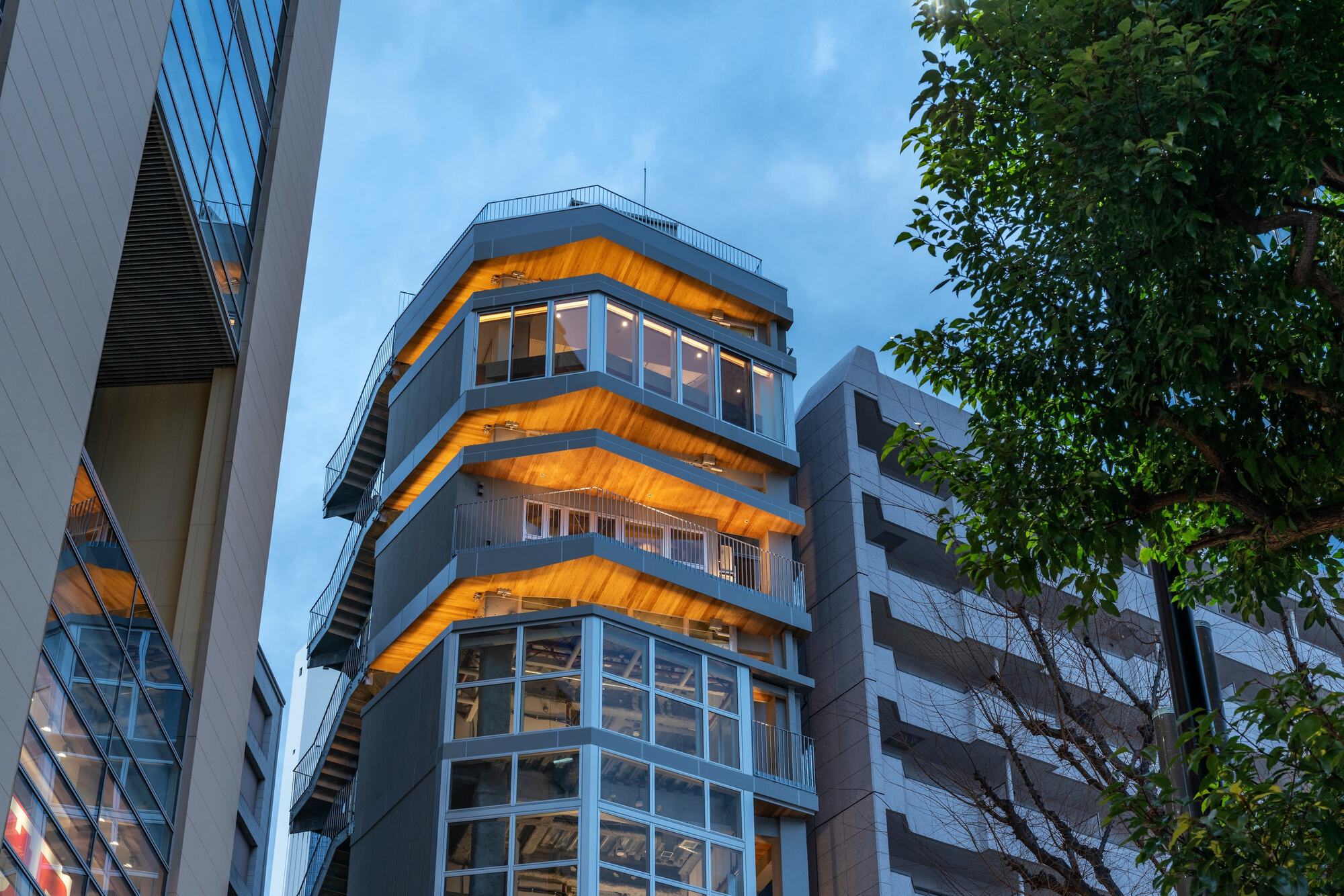 Stepped tower with glowing timber soffits and glazed corners, framed by a leafy tree against a blue evening sky