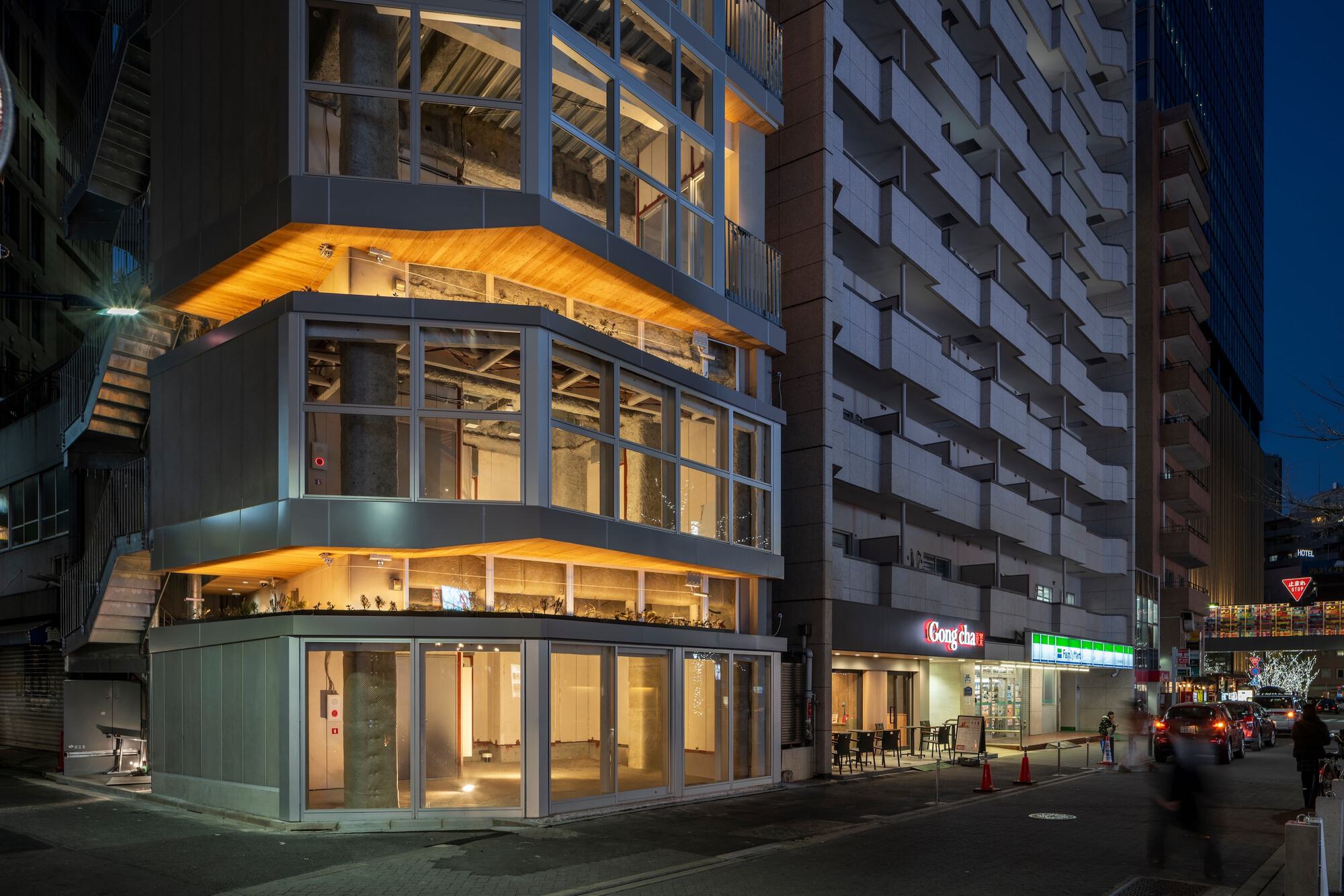 Corner of a stepped glass tower with glowing timber soffits and exposed concrete columns, seen at dusk beside a street cafe