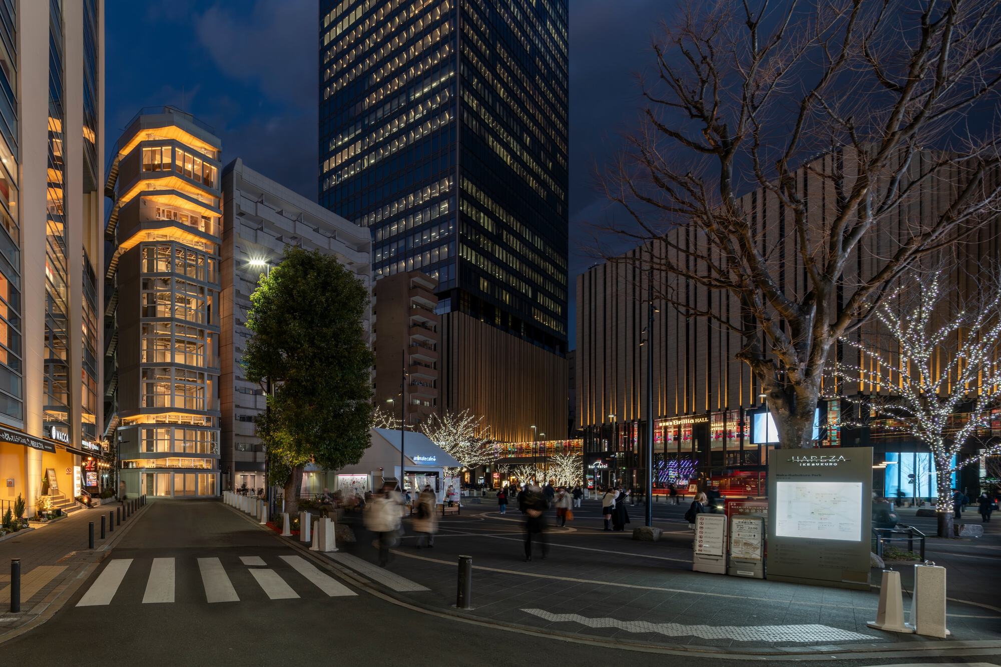 Narrow stepped tower with lit timber eaves on a plaza corner, framed by bare trees strung with lights at dusk