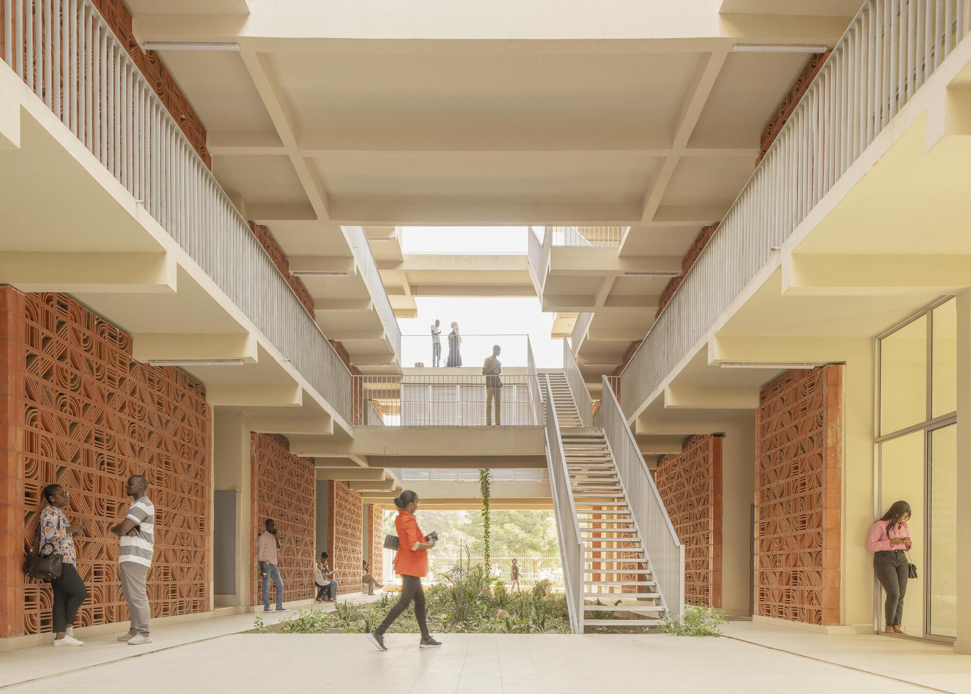 Open-air atrium with terracotta screen walls, a steel staircase and planted courtyard, with students crossing the pale floor in daylight