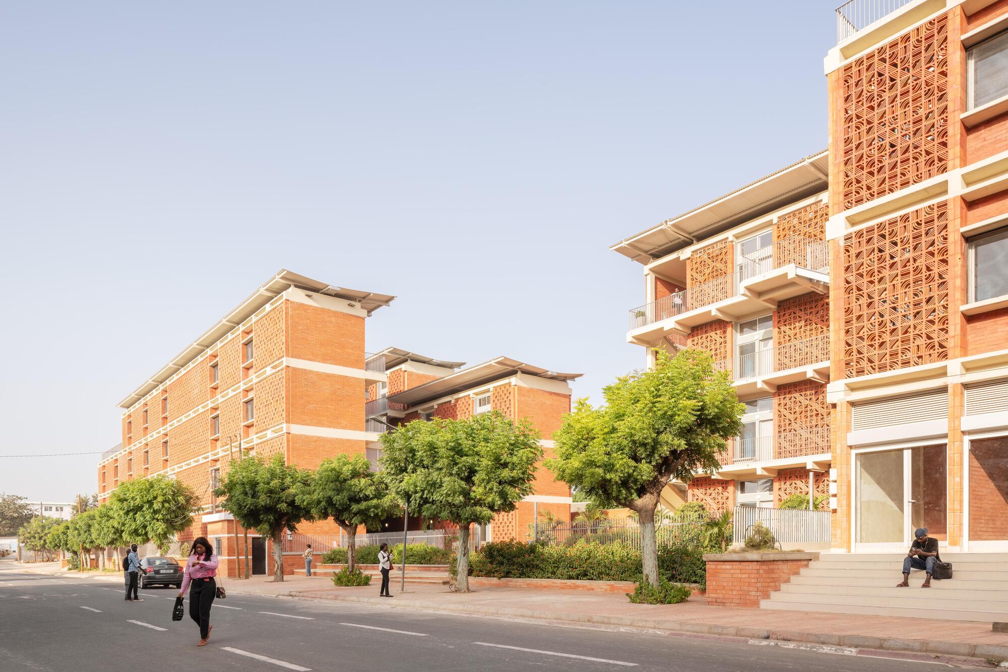 Street view of stepped brick blocks with perforated screen panels and sloped roofs, street trees in front under a hazy sky