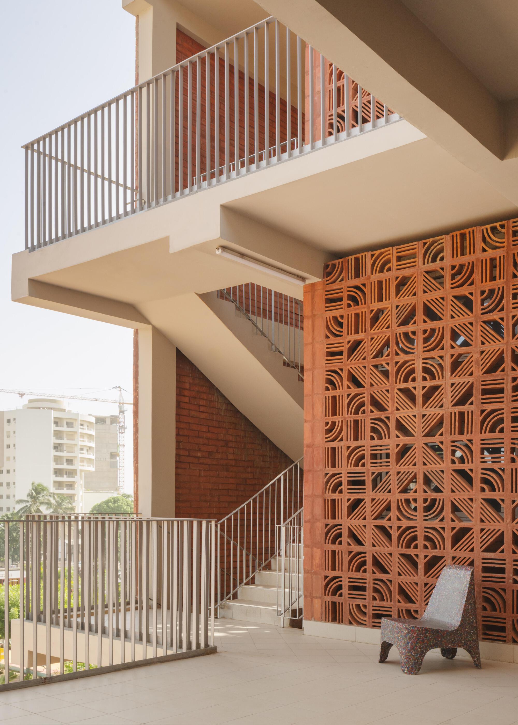 Corner of an open stairwell beside a patterned terracotta screen wall, with a recycled-material chair on the landing in soft daylight