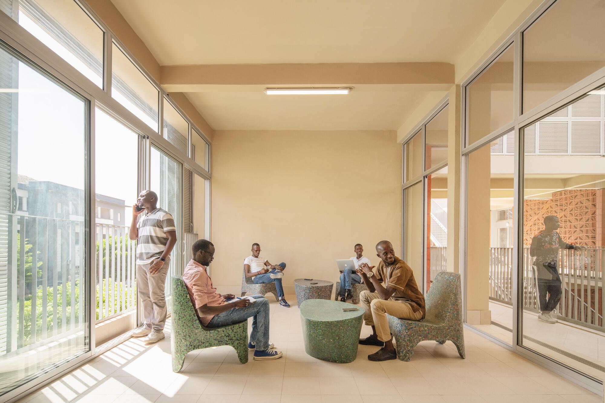 Glazed lounge with speckled green chairs and low tables where four men talk and work, sunlight falling across the floor