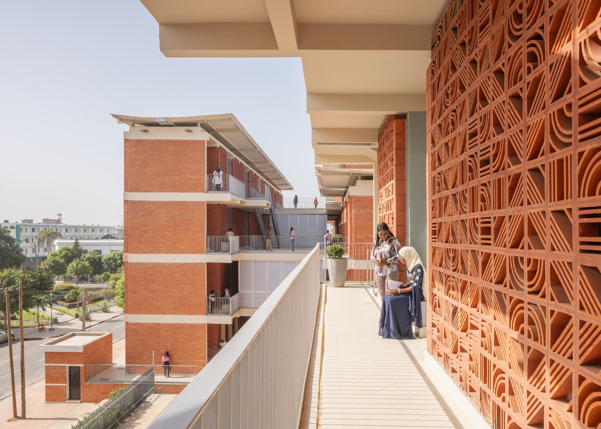 Upper walkway running beside a terracotta screen wall, with two women talking near a planter and a brick block beyond