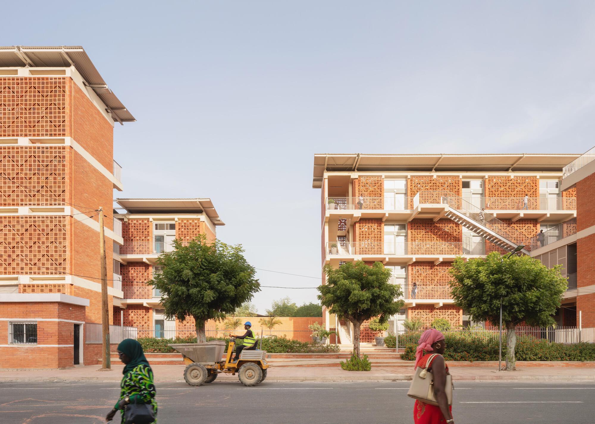 Street view of brick lattice facades and stair-linked balconies, with a small dumper and pedestrians passing in front of young trees