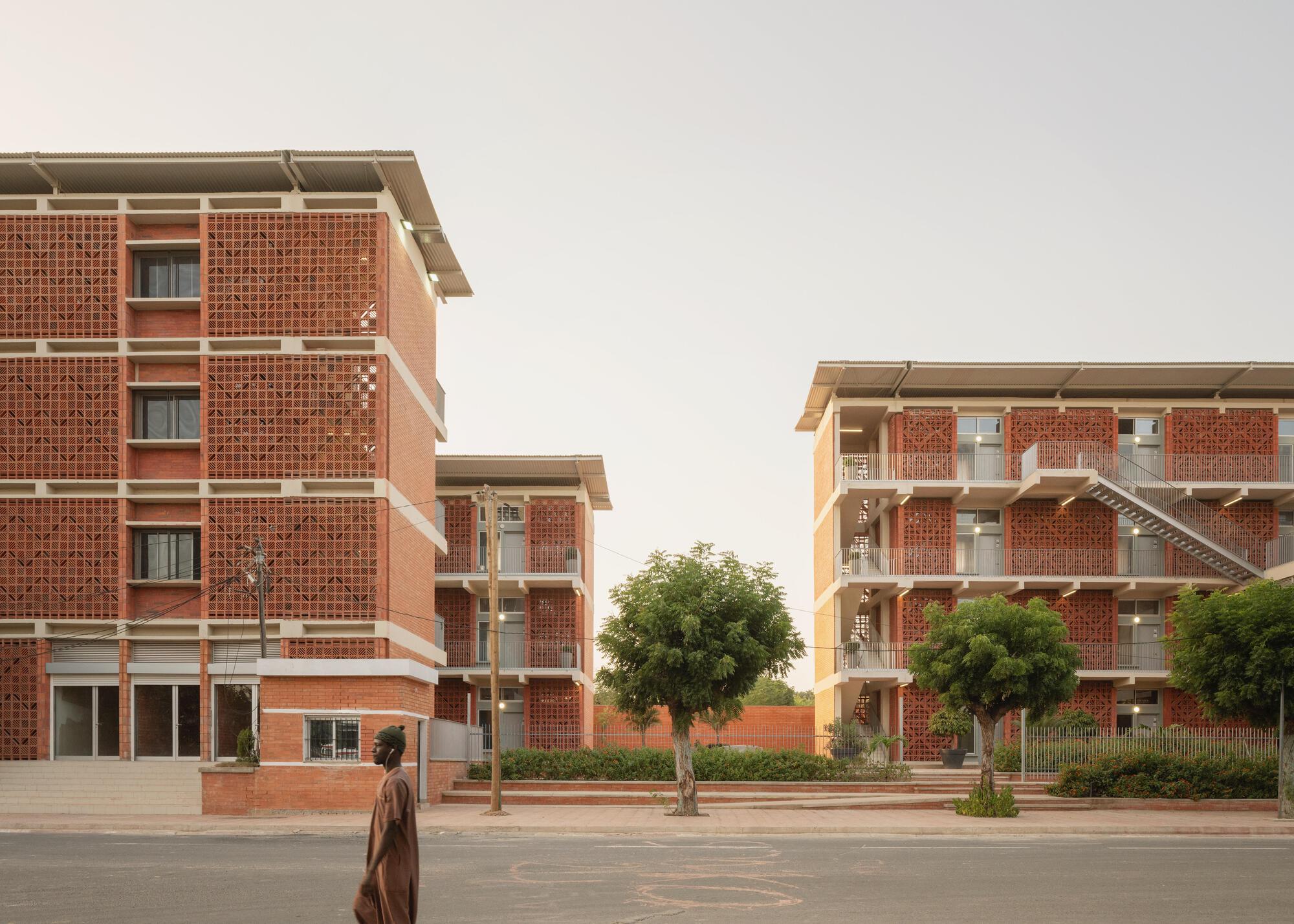 Two brick blocks with perforated screen walls and projecting roofs, a man in a long robe walking past at dusk
