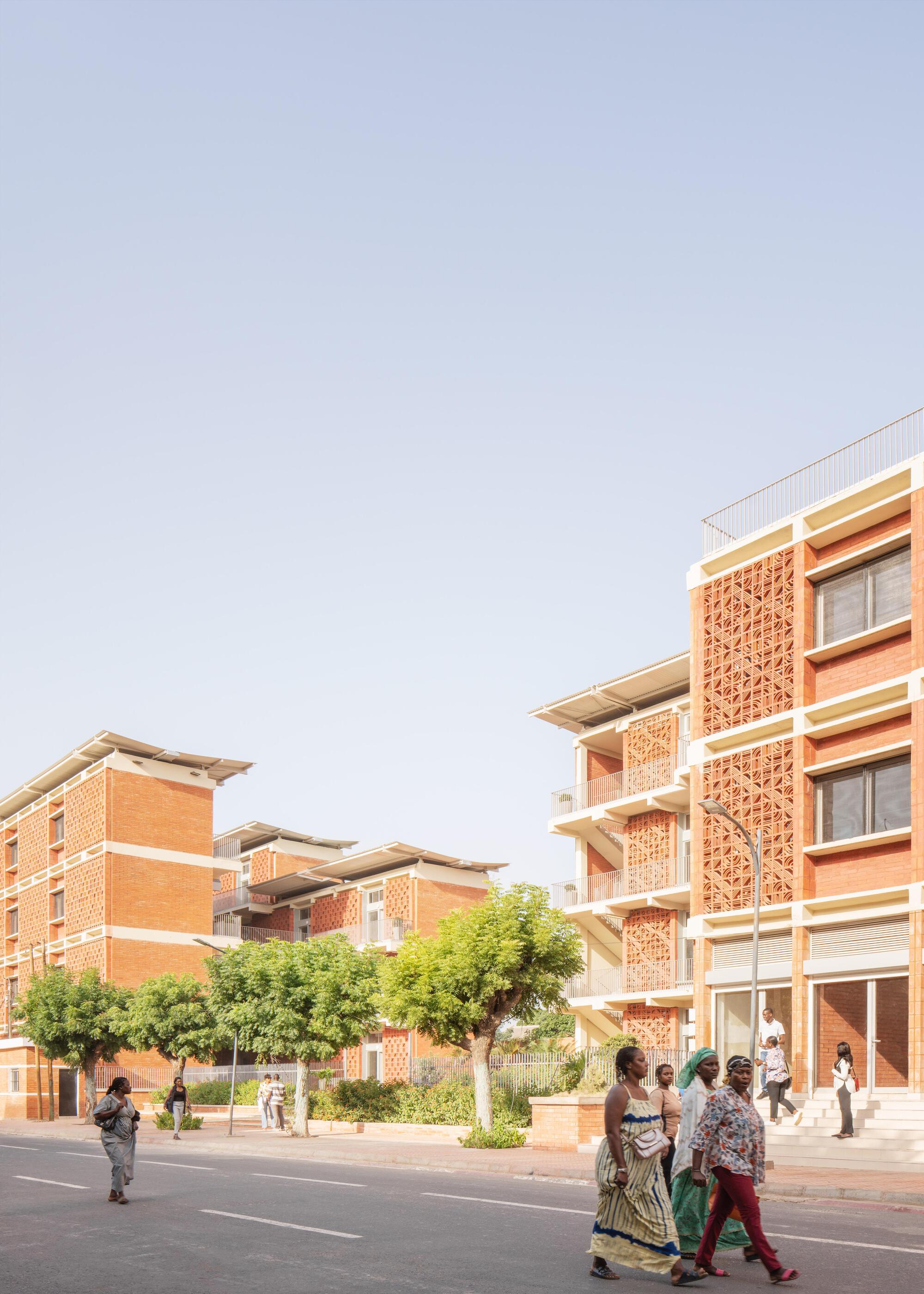 Vertical view of the brick lattice facade and white banded balconies, with women in colorful dresses crossing the street in front