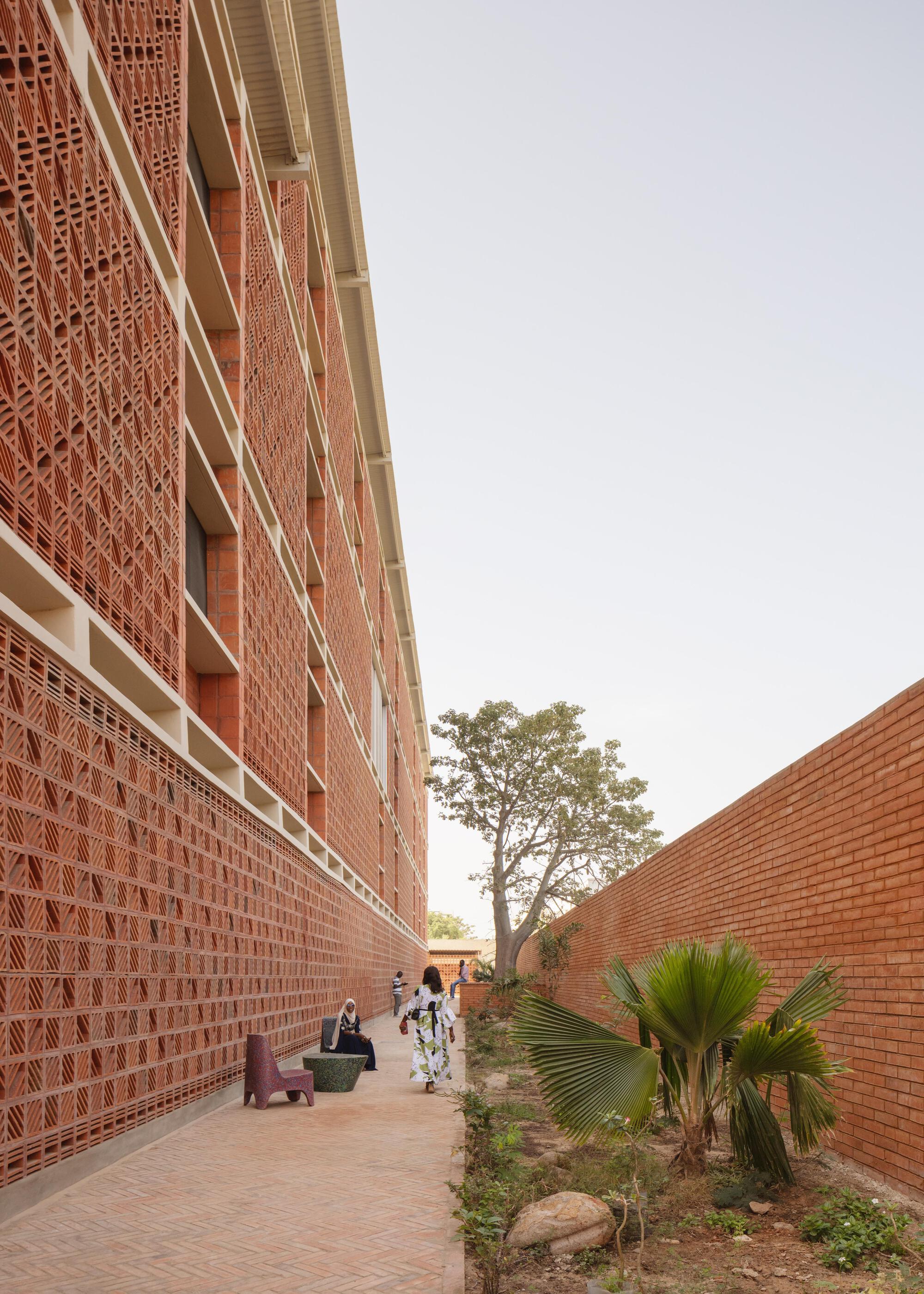 Narrow brick-paved passage along a tall lattice brick wall, with a woman walking toward a baobab-like tree and a fan palm