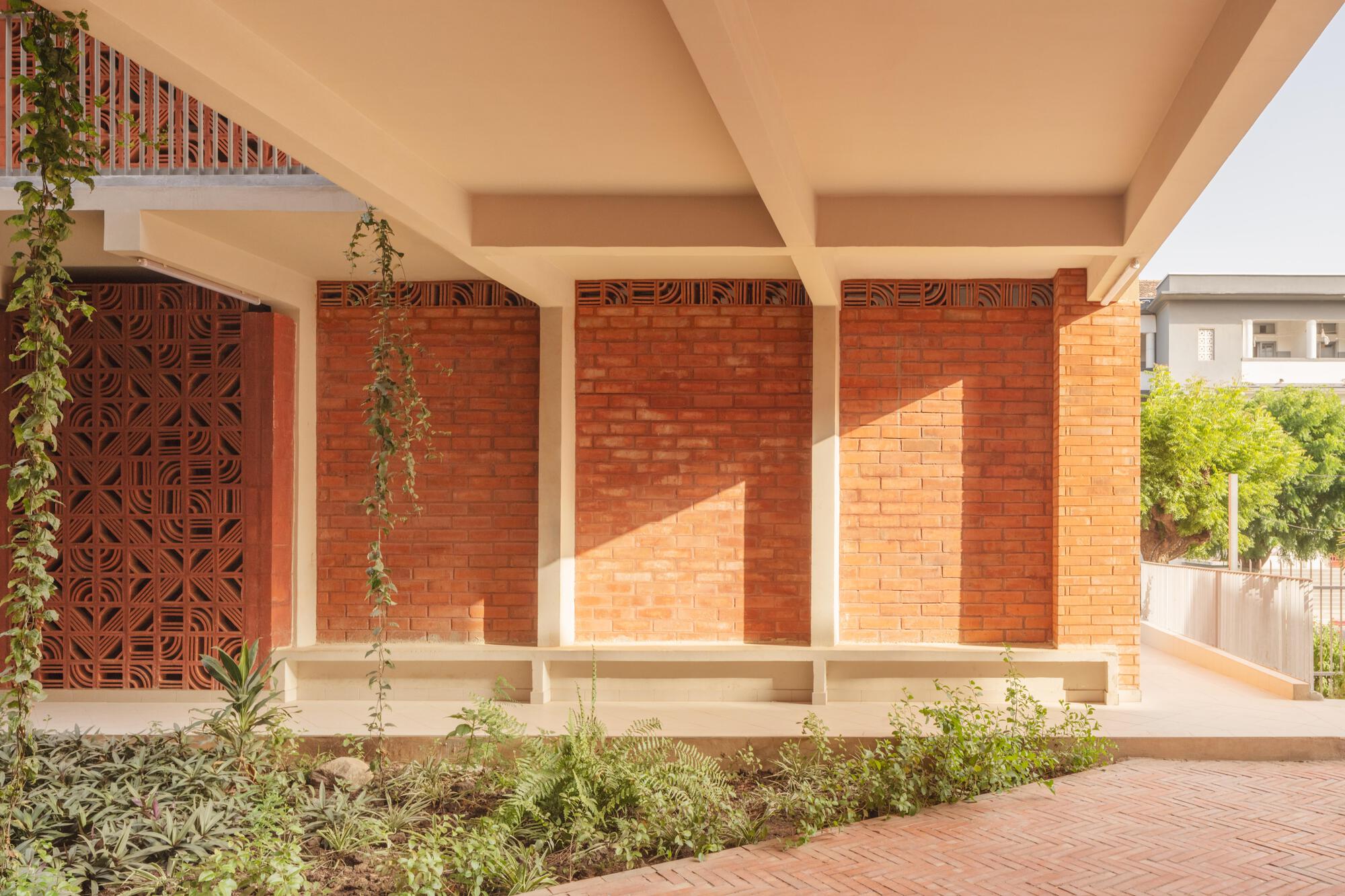 Covered walkway with brick infill panels, concrete beams and a perforated screen door, with vines hanging over a planted bed in afternoon sun