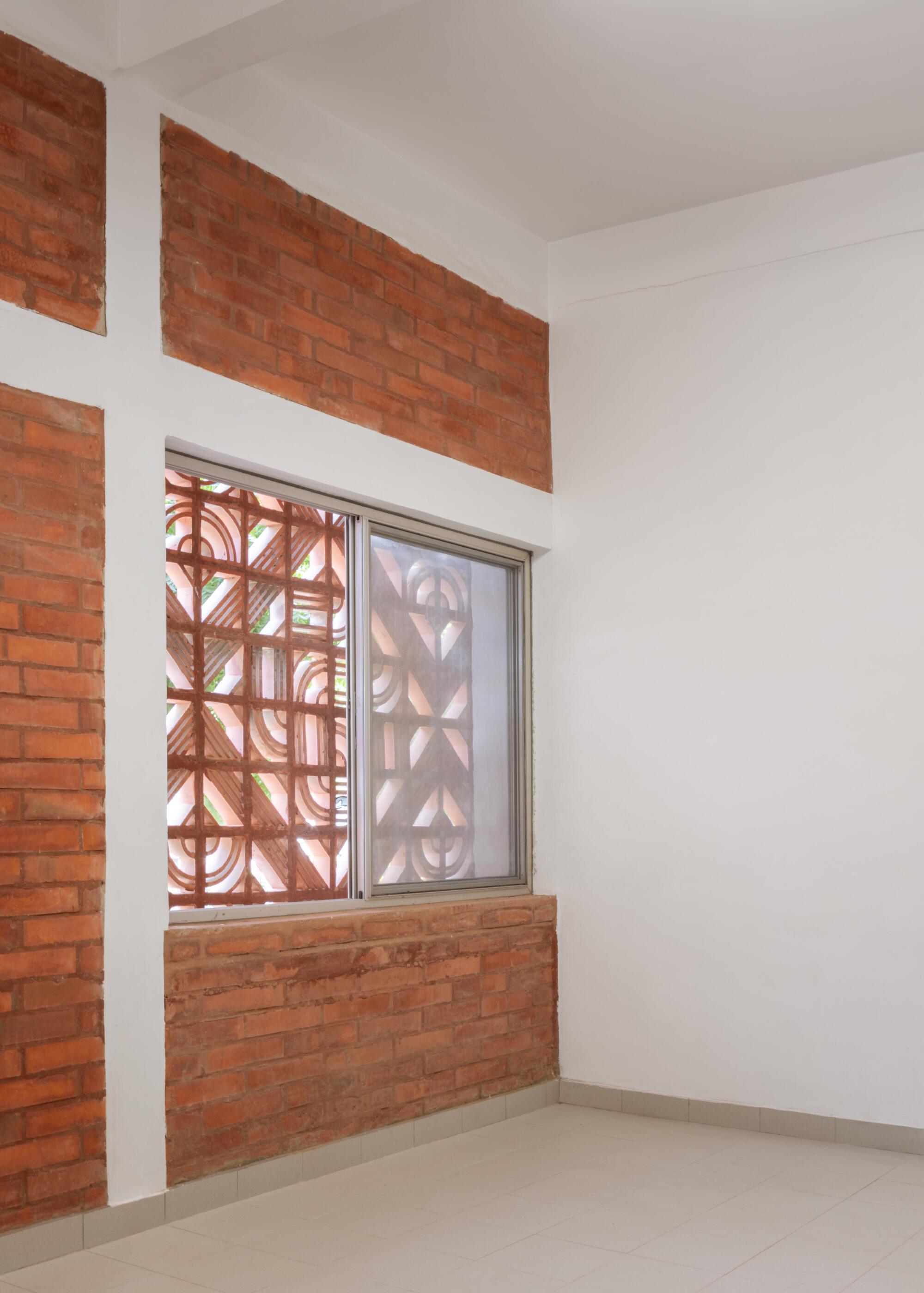 Corner of a white room with exposed brick bands and a sliding window framing a patterned terracotta screen in daylight