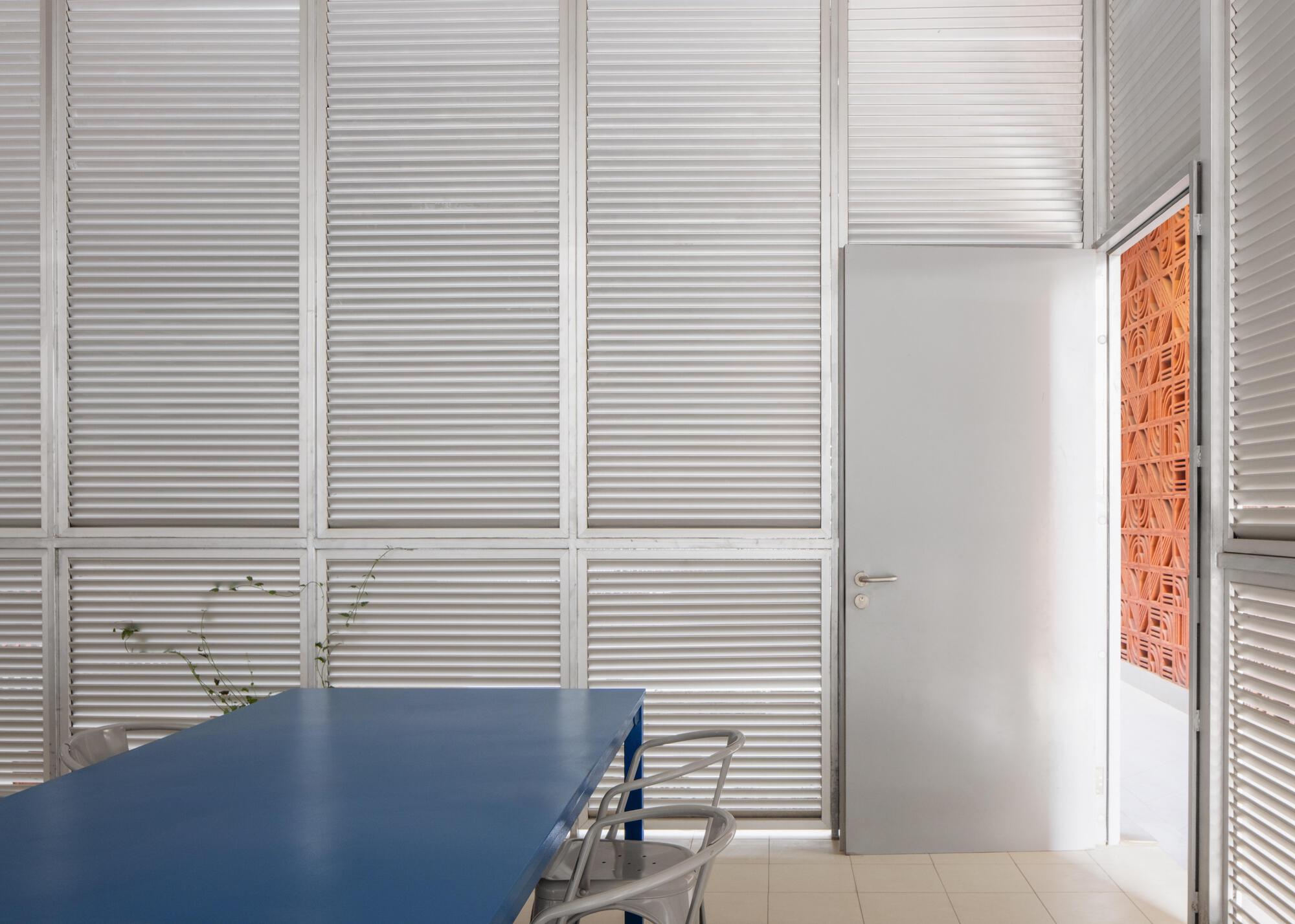 Meeting room walled in white louvers, with a blue table and metal chairs beside an open door onto a terracotta screen