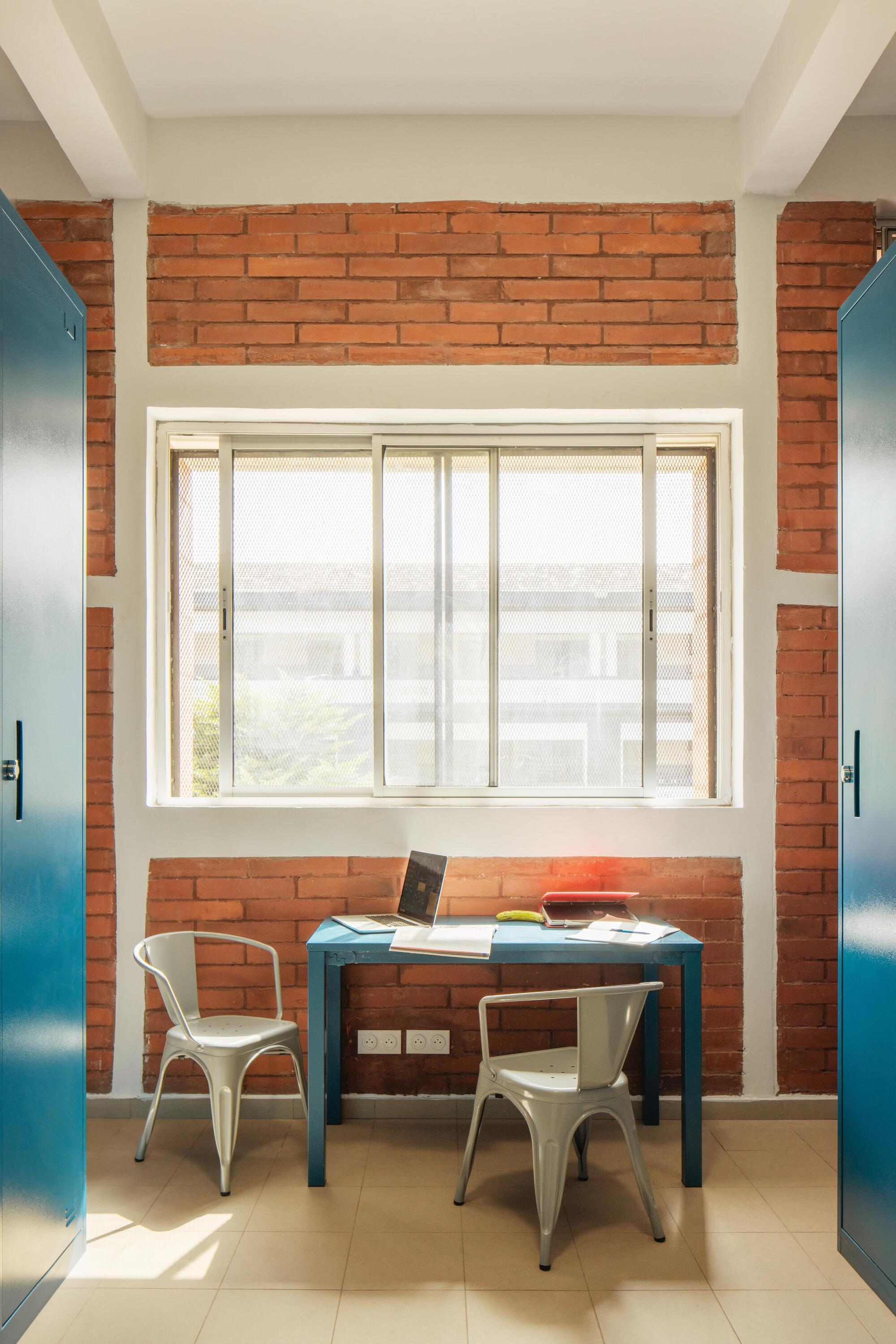 Study nook with a blue desk, two metal chairs and brick bands around a sliding window, flanked by blue wardrobes in sunlight