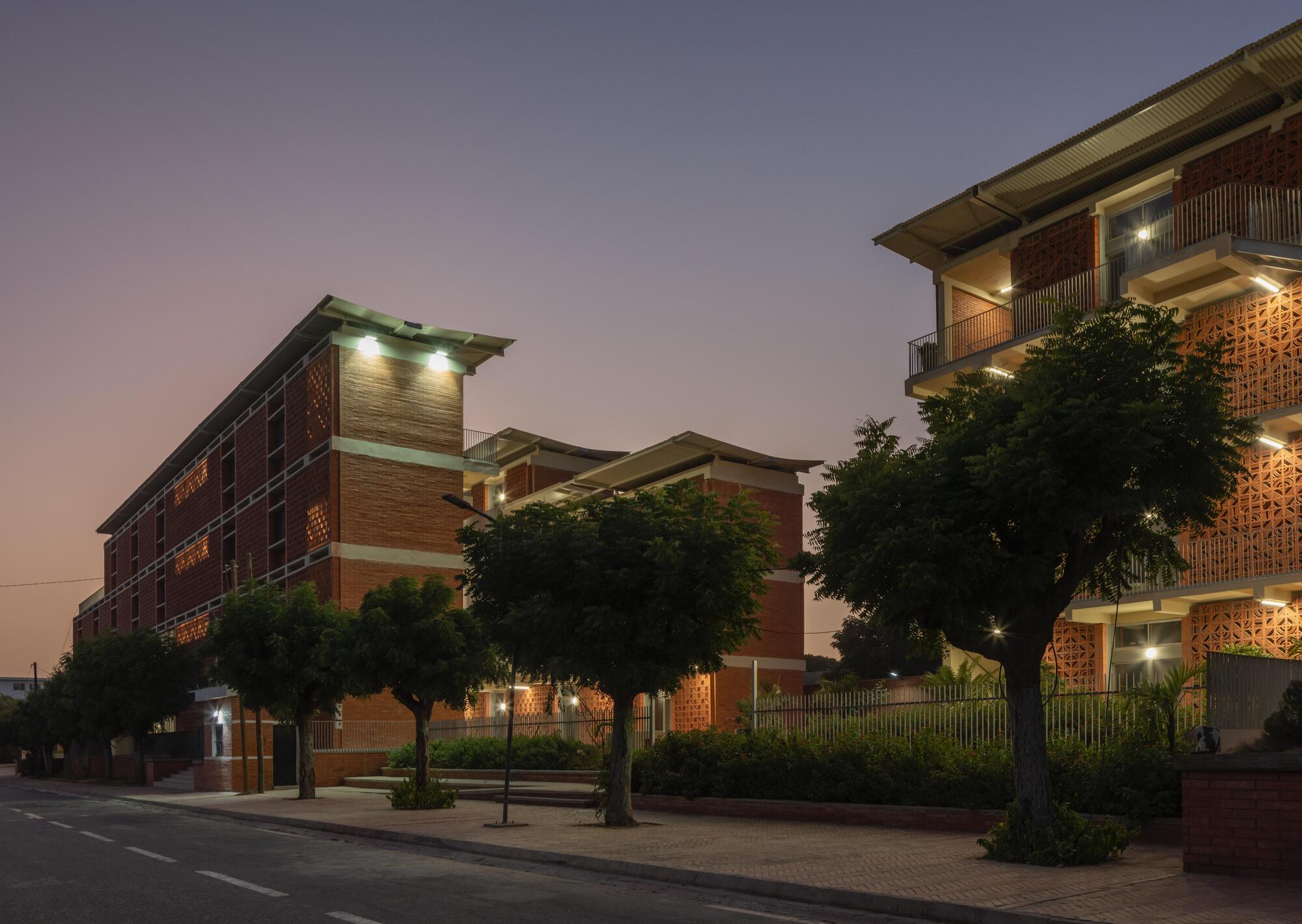 Brick dormitory blocks with perforated screen walls and lit balconies behind street trees at dusk