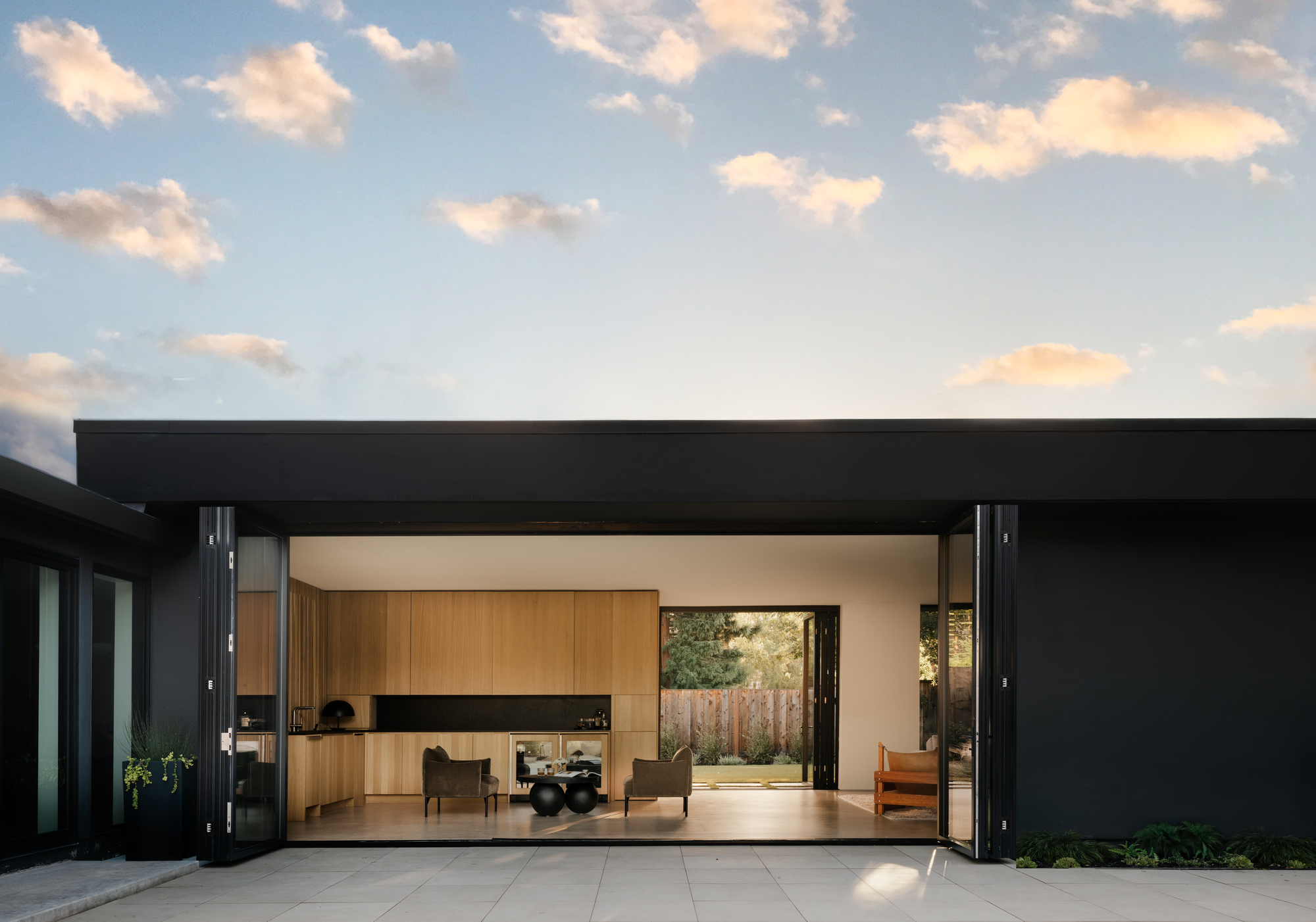 Black single-storey pavilion with folding glass doors opened to a kitchen and garden, under a dusk sky with pink-lit clouds