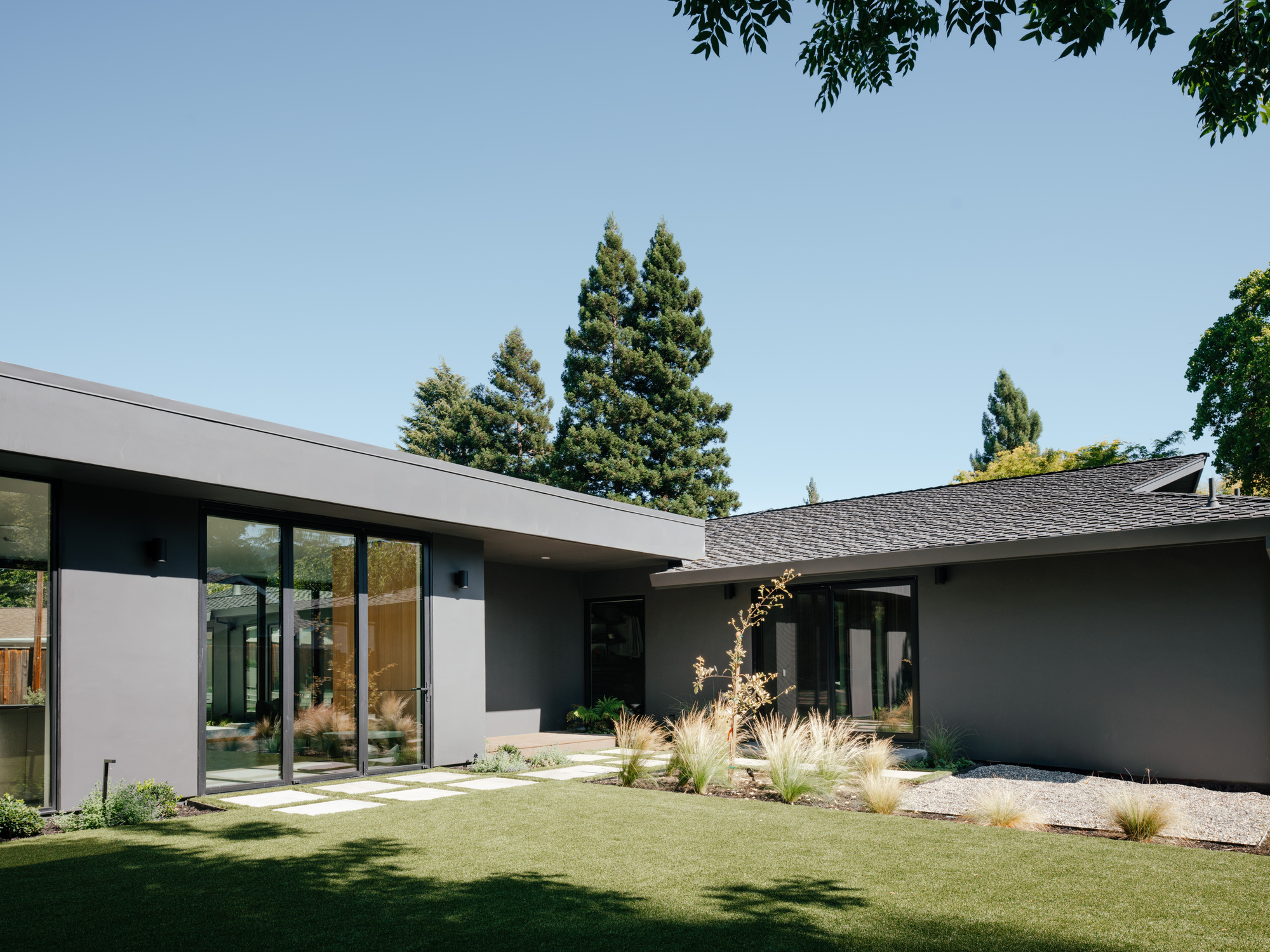 Grey stucco courtyard house with flat and pitched roofs, glazed walls and grasses planted along a stepping-stone path in sun