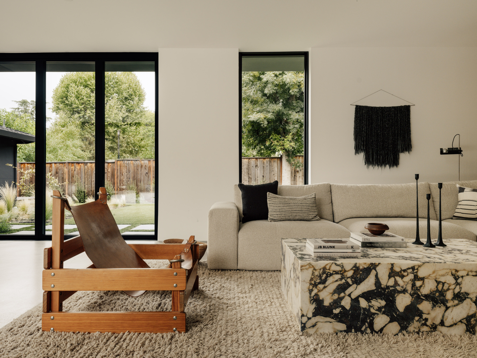 Living area with a leather and timber chair, marble coffee table and woven wall hanging, tree-filled garden visible through black-framed glass