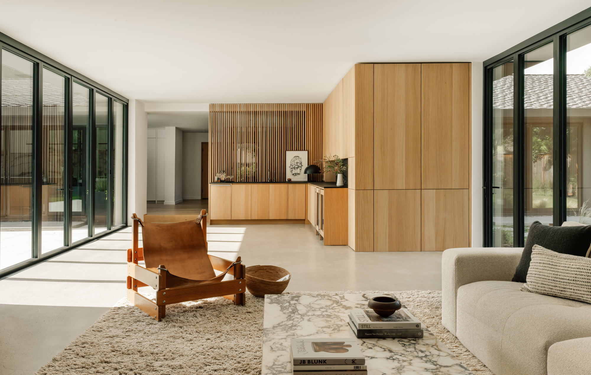 Long living space with oak slat wall, tall cabinet block and floor-to-ceiling glazing, afternoon light spilling over the polished concrete floor
