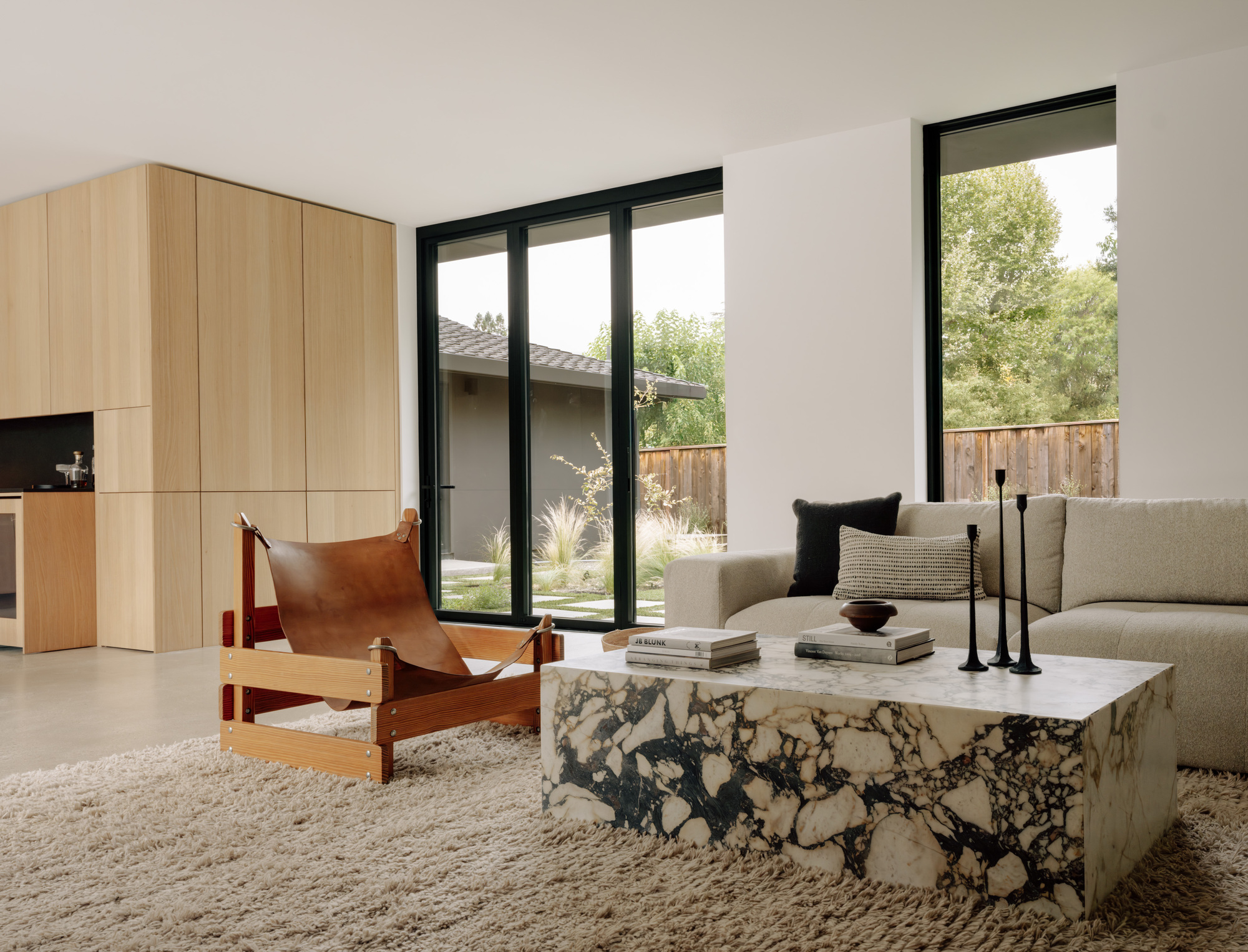 Living area with a veined marble coffee table, leather sling chair and oak cabinetry beside black-framed glass doors onto a garden