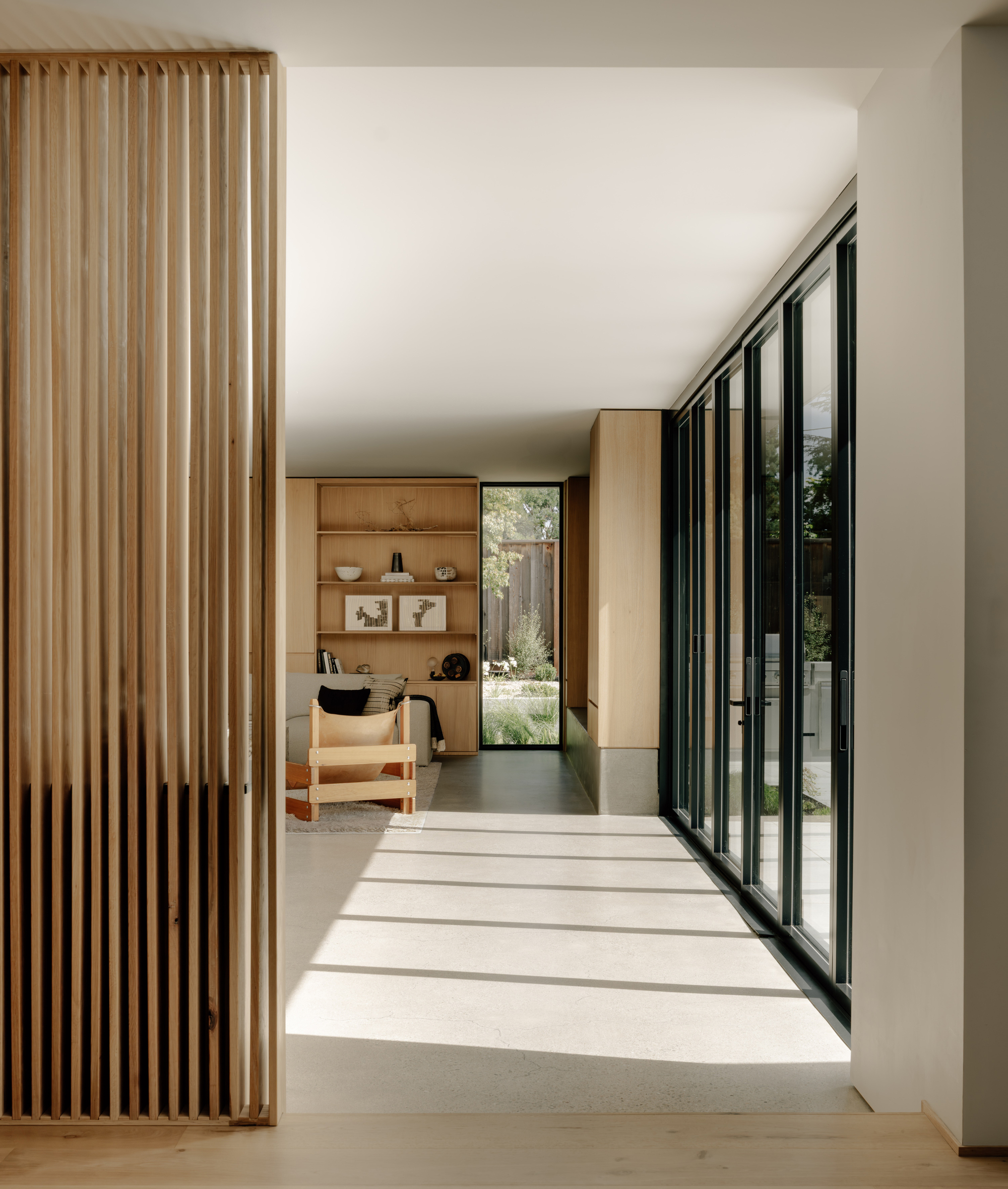 Hallway past an oak slat screen, with sunlight striping the concrete floor beside a wall of folding glass doors