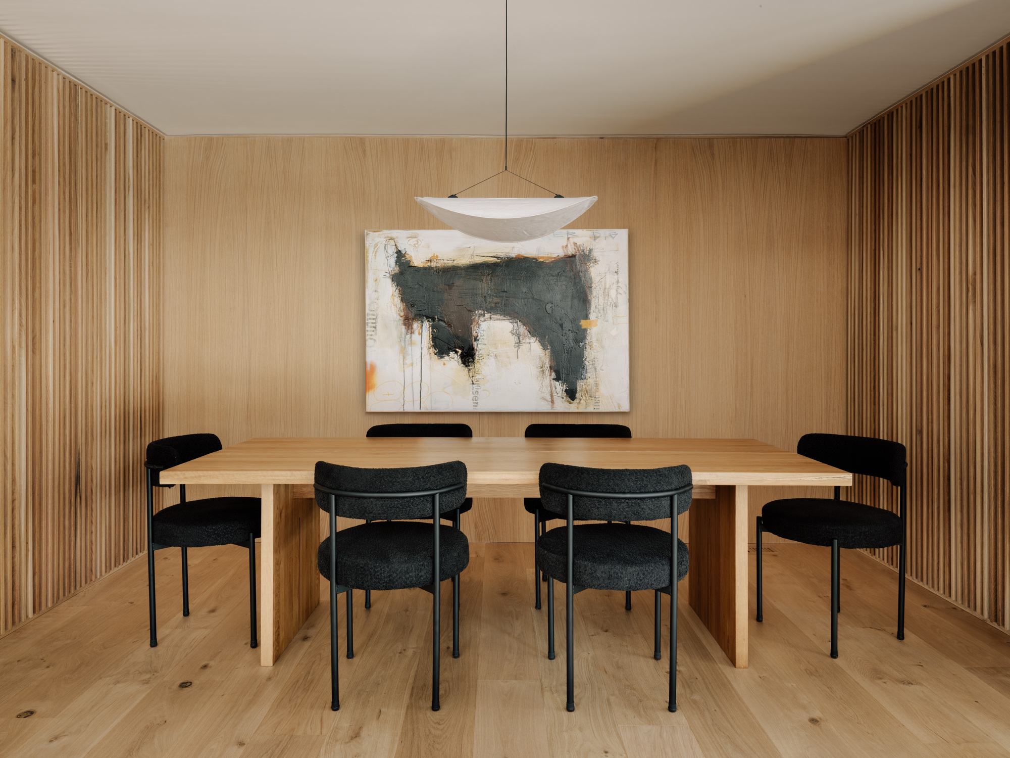 Dining room with a long oak table, black boucle chairs and a pendant lamp against vertical oak slat walls