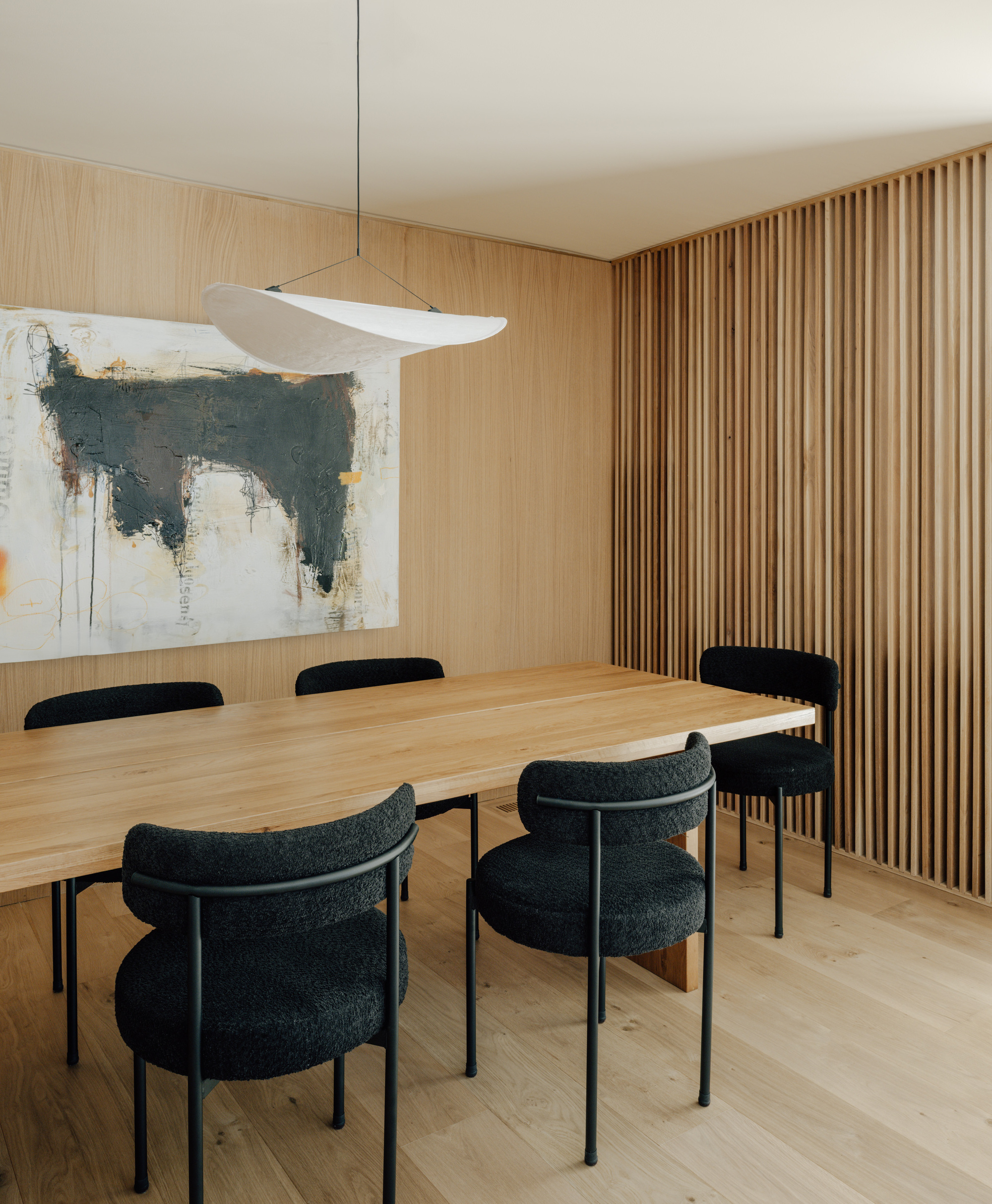 Angled view of the oak dining table, black chairs and abstract painting beside a wall of vertical timber slats