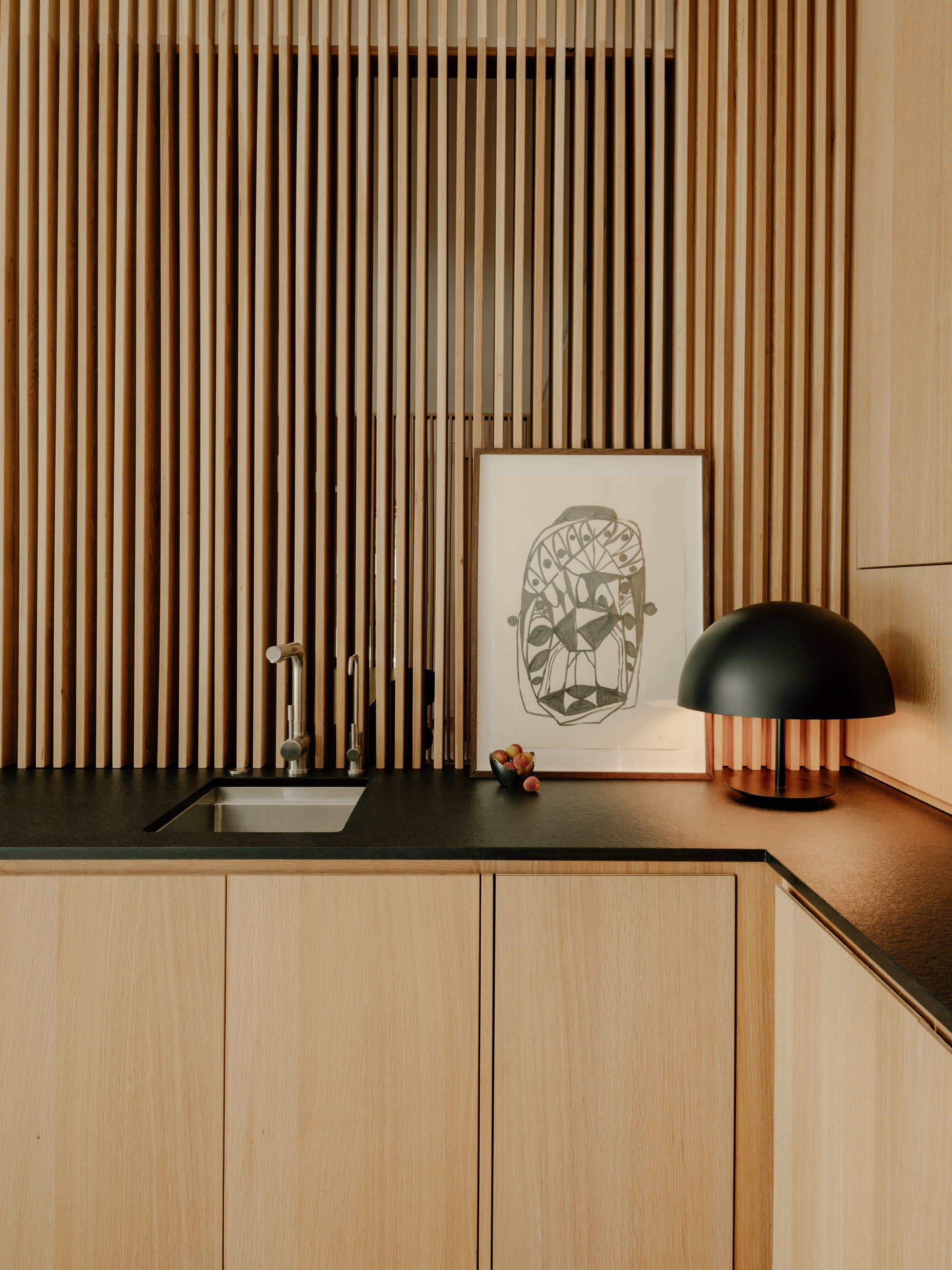 Kitchen corner with a small steel sink, black countertop and framed print against vertical oak slats lit by a dome lamp