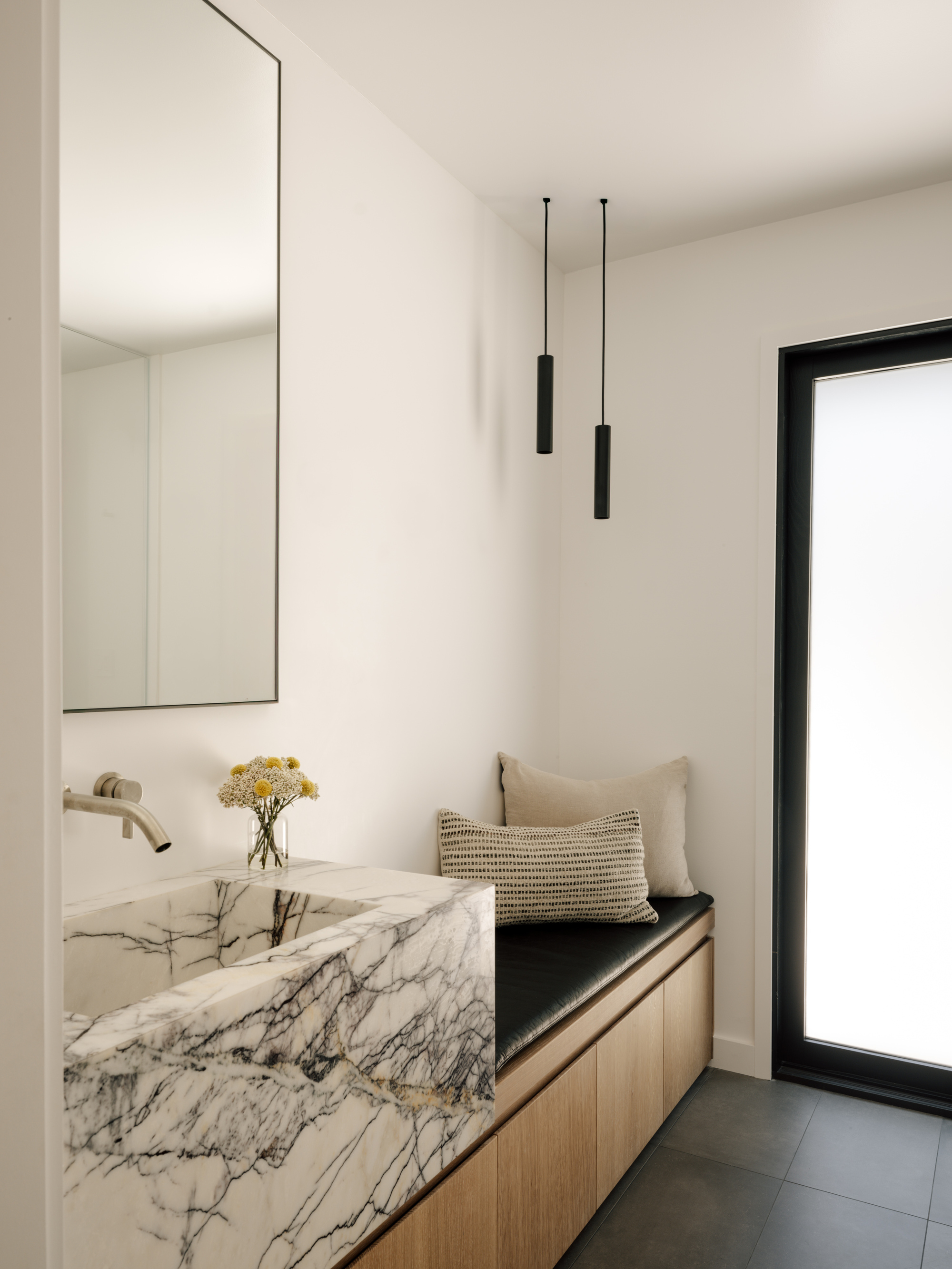 Carved marble sink and built-in leather bench with oak drawers beside a frosted glass door, two black pendants overhead