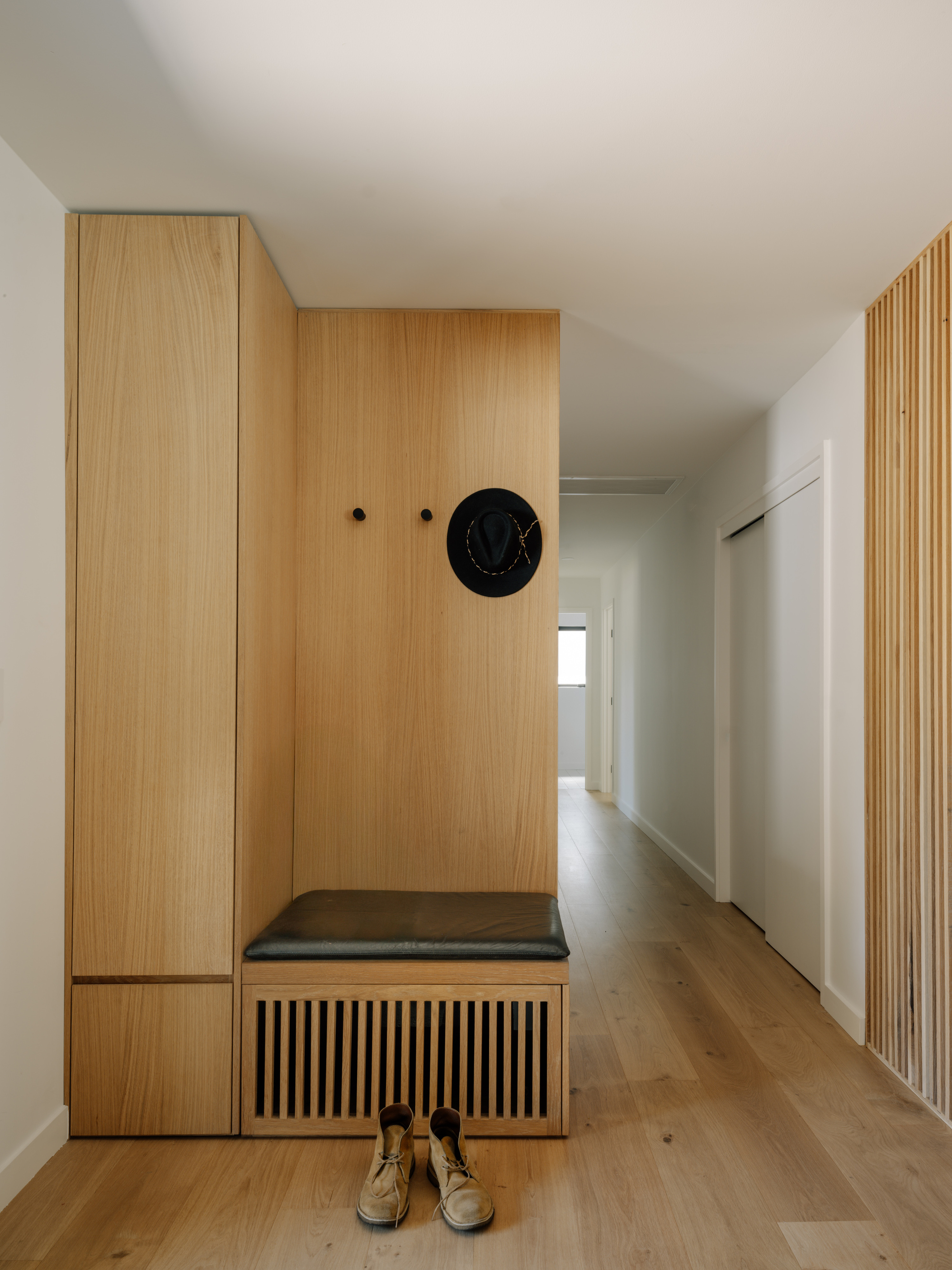 Entry nook with oak storage wall, leather bench over a slatted vent, a hat on a peg and suede boots below