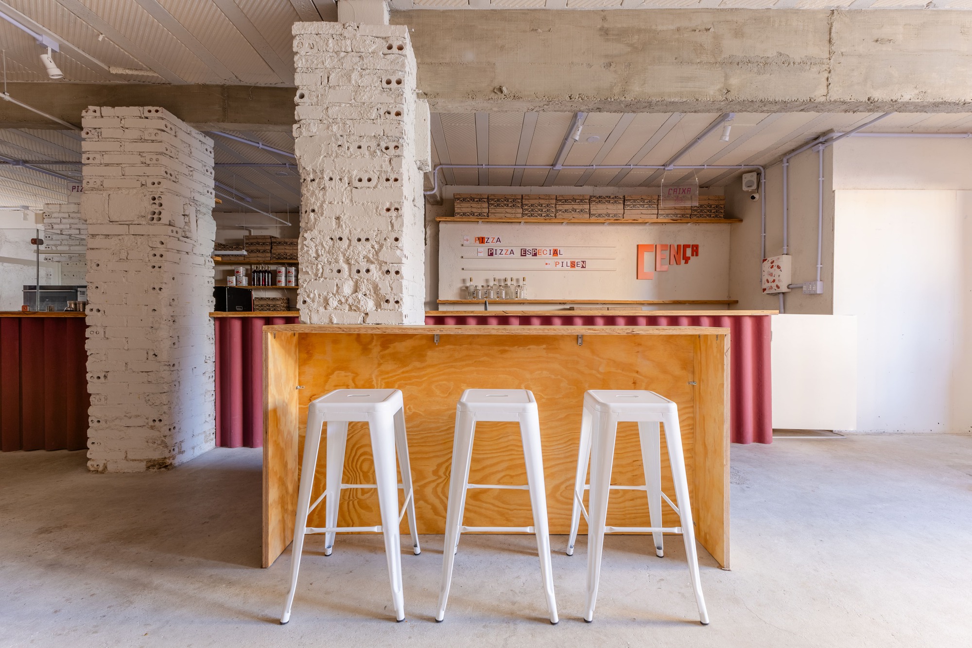 Plywood bar counter with three white metal stools in front of a painted brick pillar and a menu wall