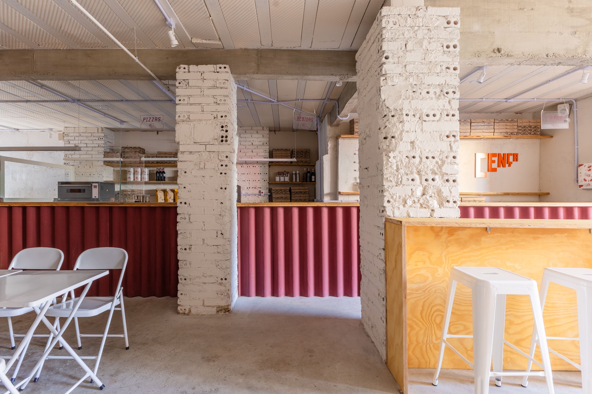 View between painted brick pillars toward the red corrugated counter and the plywood bar with white stools beyond