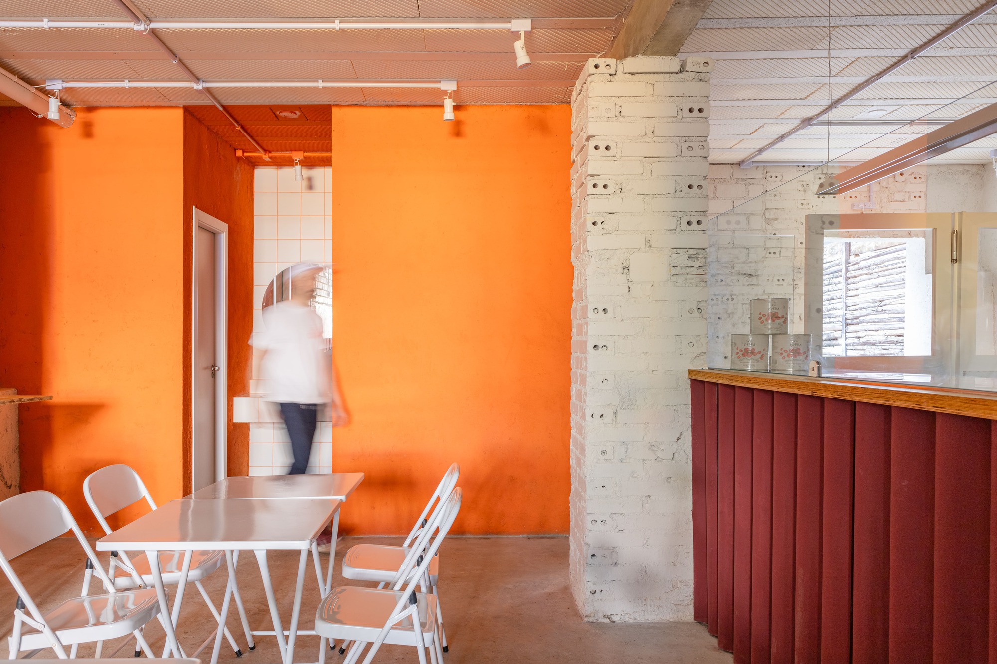 Orange painted walls and a blurred figure walking past white folding tables, with a red ribbed counter at right