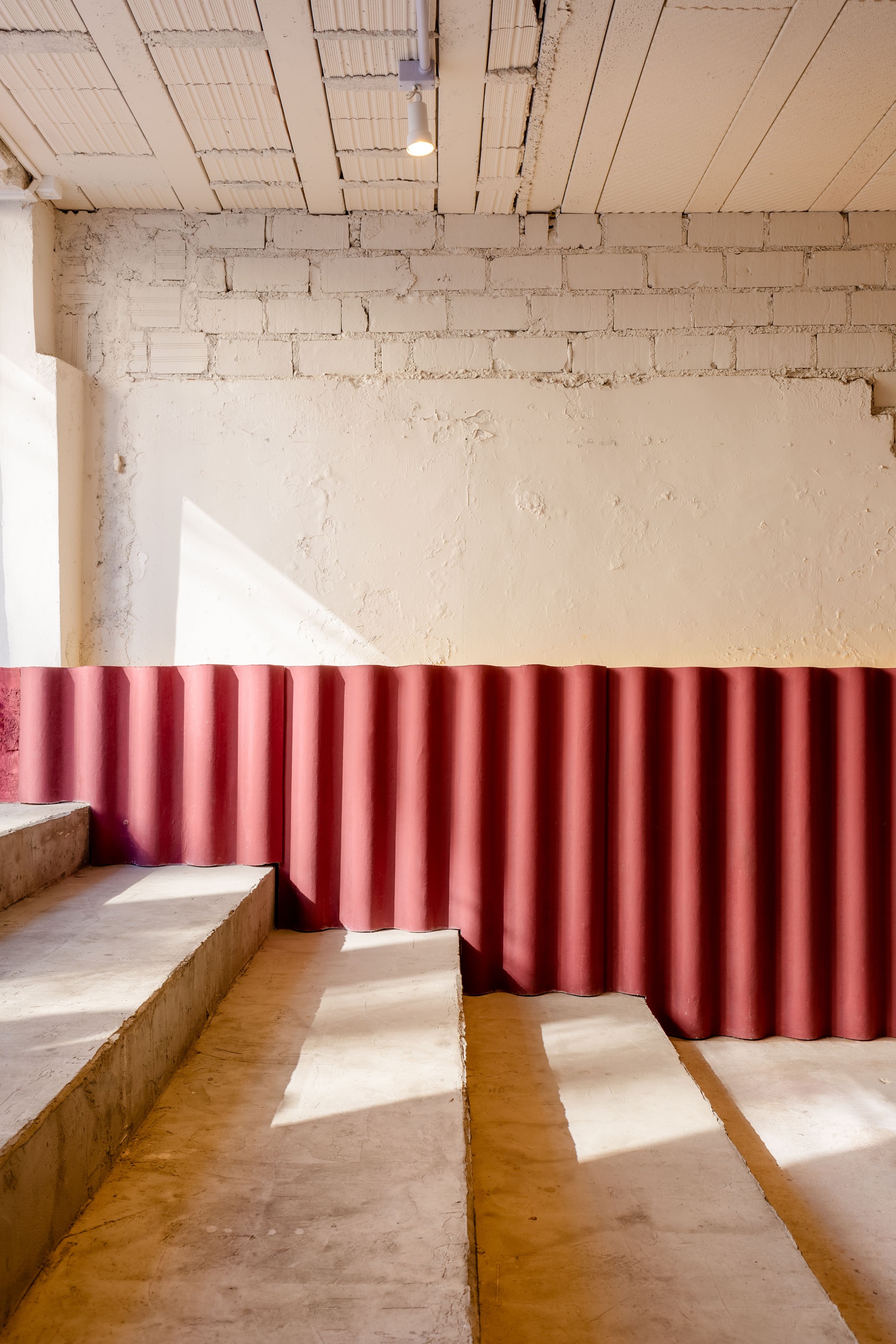 Concrete steps beside a red corrugated wall panel under a whitewashed brick wall, with sunlight casting diagonal shadows
