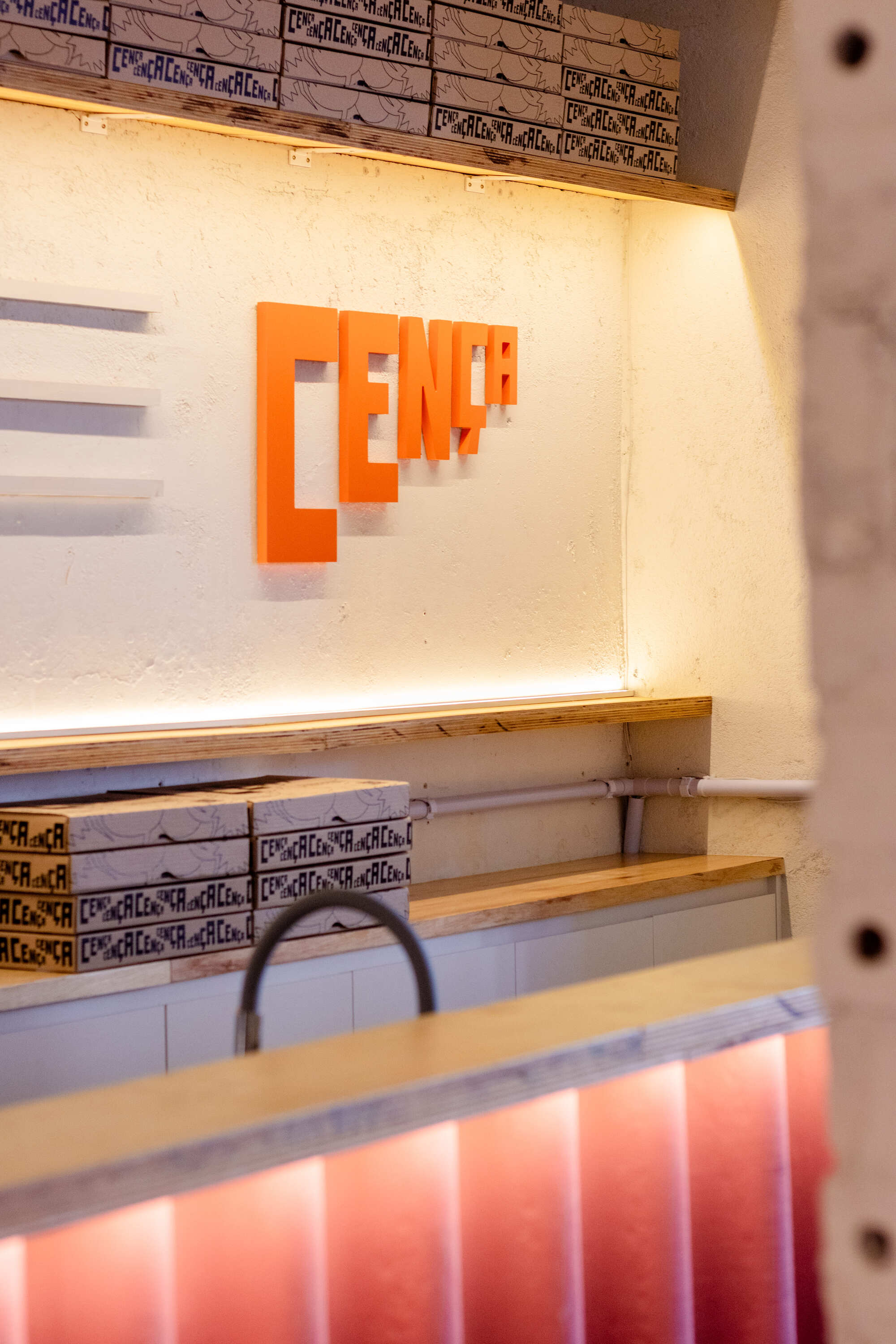 Orange wall lettering above a plywood counter lit from below, with stacked pizza boxes on the shelves in warm light