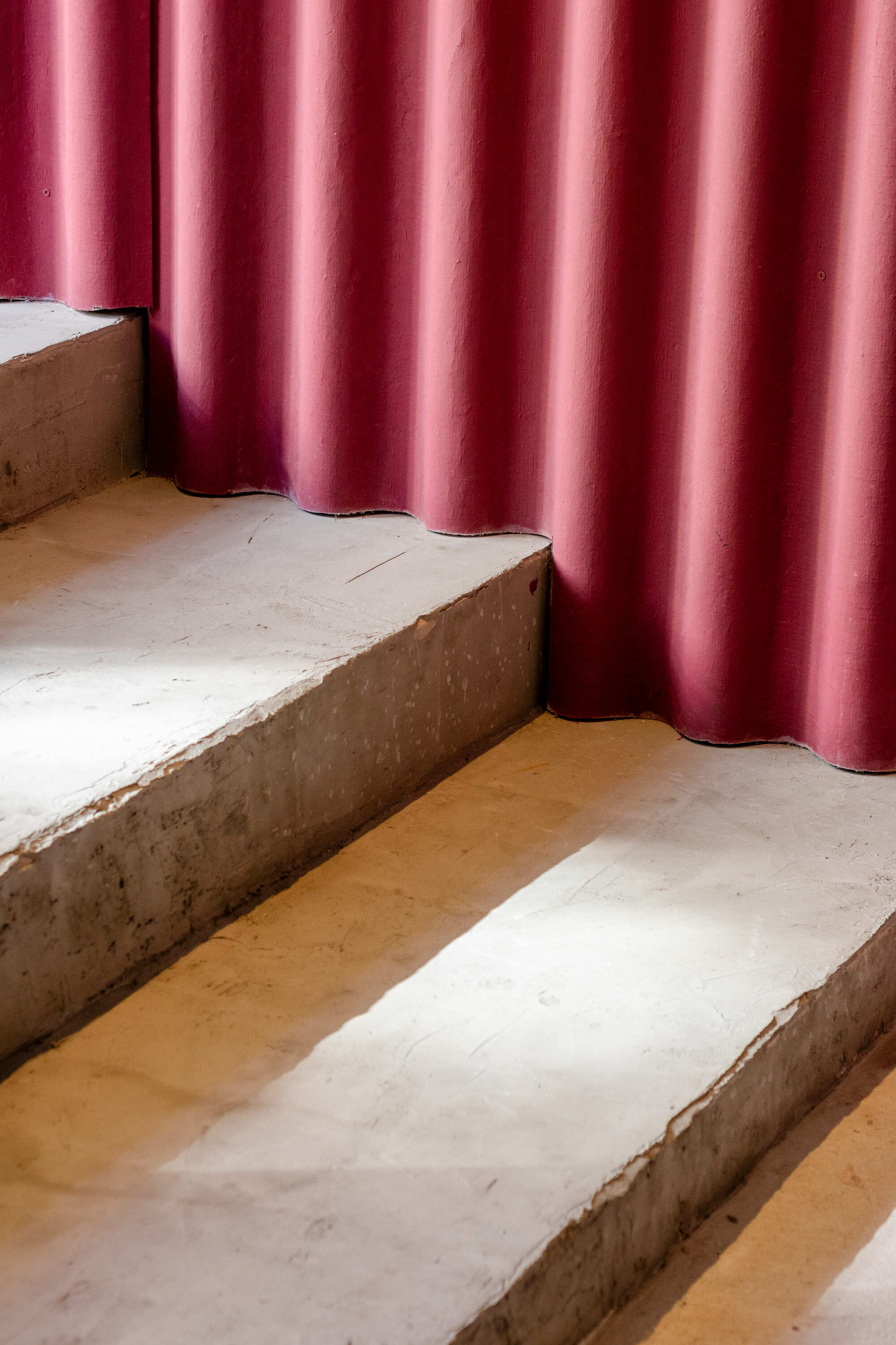 Rough concrete steps meeting a wavy maroon corrugated wall, with strips of sunlight falling across the treads