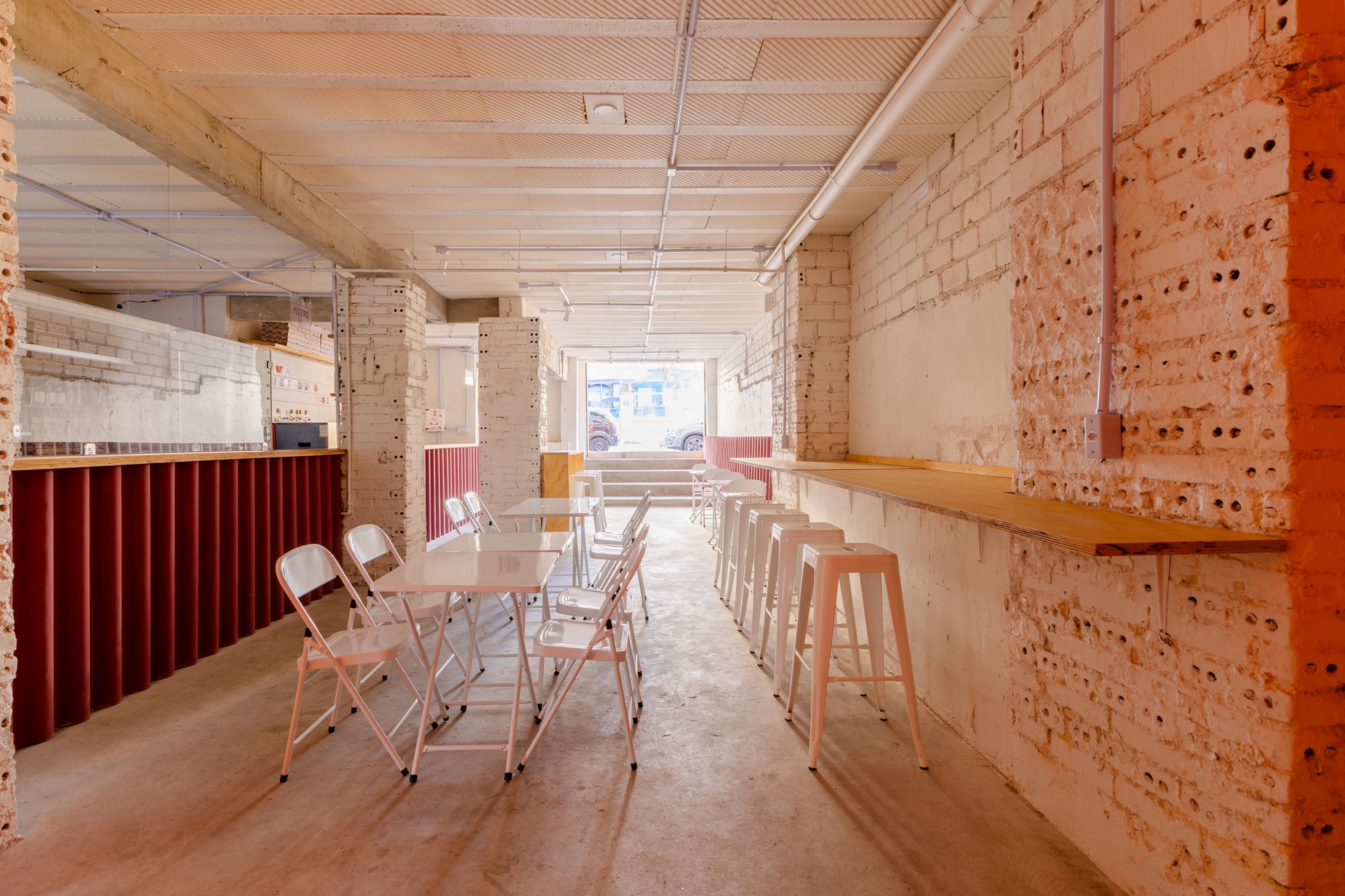 Long narrow dining room with folding chairs, white stools at a timber ledge and a maroon corrugated counter, daylight at the far entrance