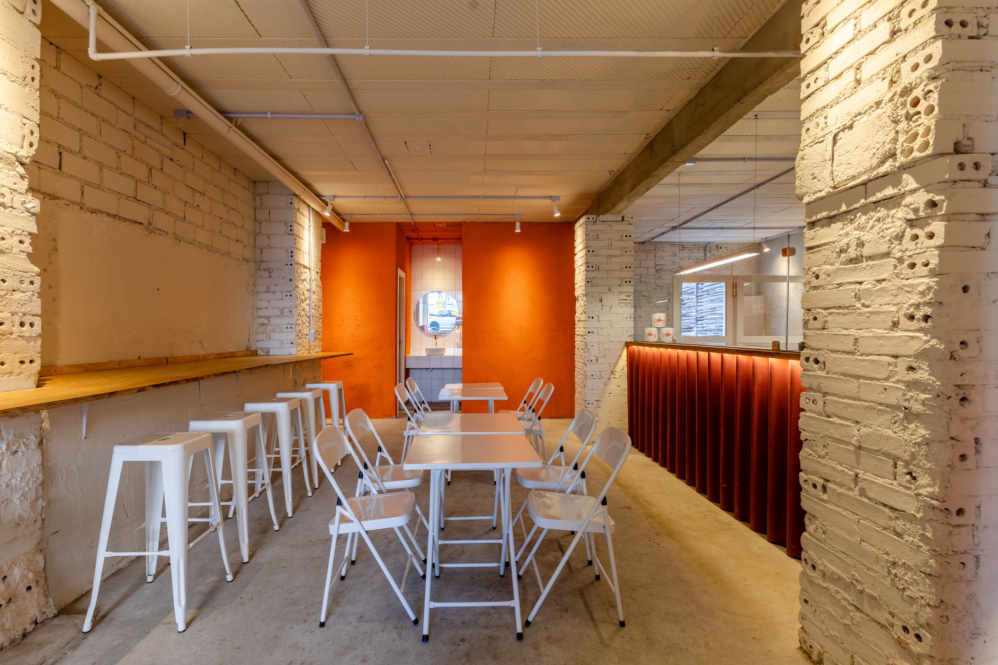 Dining room with white folding chairs and tables, tall stools at a wall ledge, and an orange wall at the far end