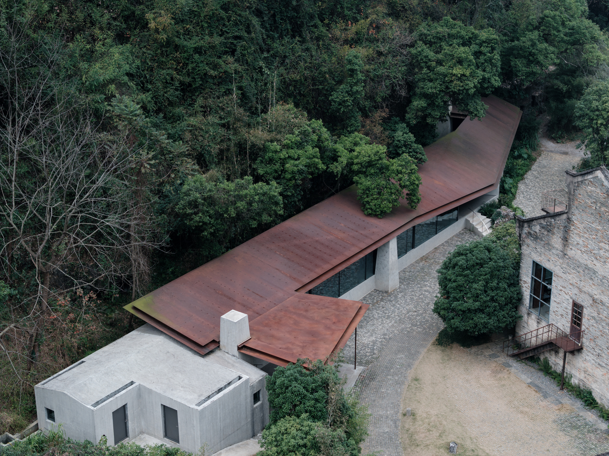 Aerial view of the folded weathering-steel roof running along a wooded slope beside a concrete volume and old stone building