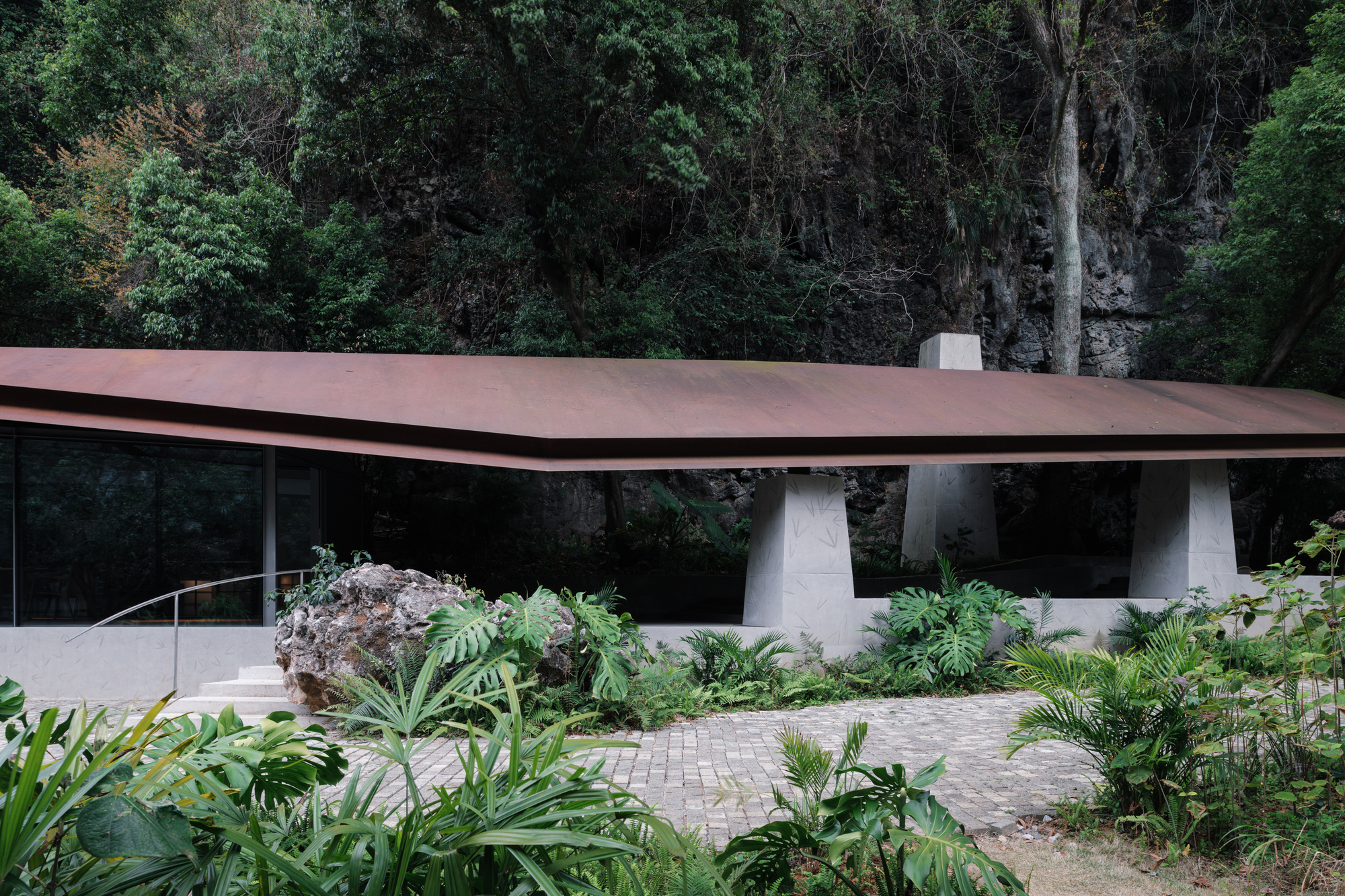 Low rusted steel roof resting on tapered concrete piers, with a boulder and tropical plants in the foreground under dense forest