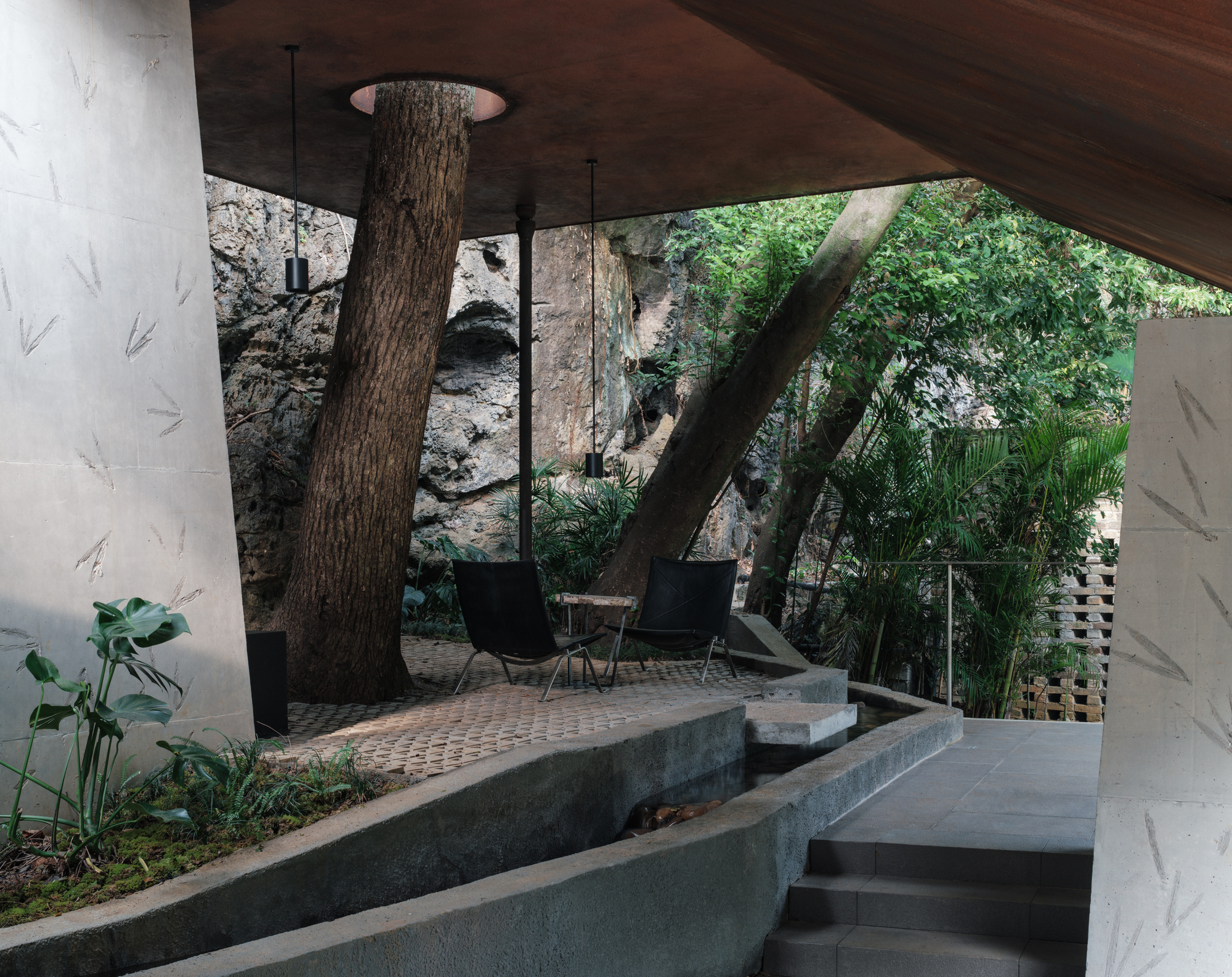 Covered terrace where a living tree trunk passes through the roof, beside a narrow water channel and leaf-imprinted concrete piers