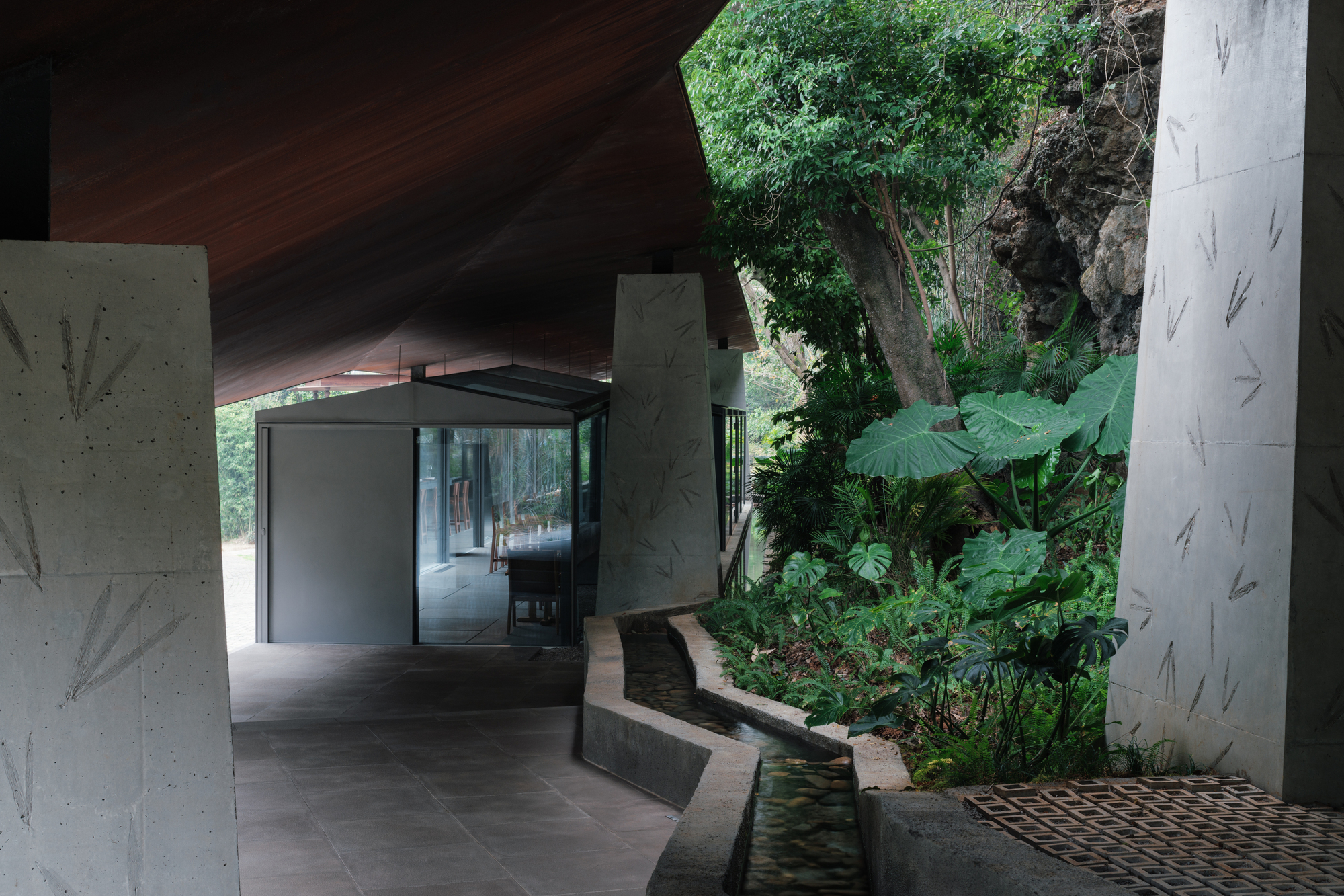 Shaded walkway under the timber-lined roof, with a stone-lined water channel and large-leaf plants beside leaf-patterned concrete piers