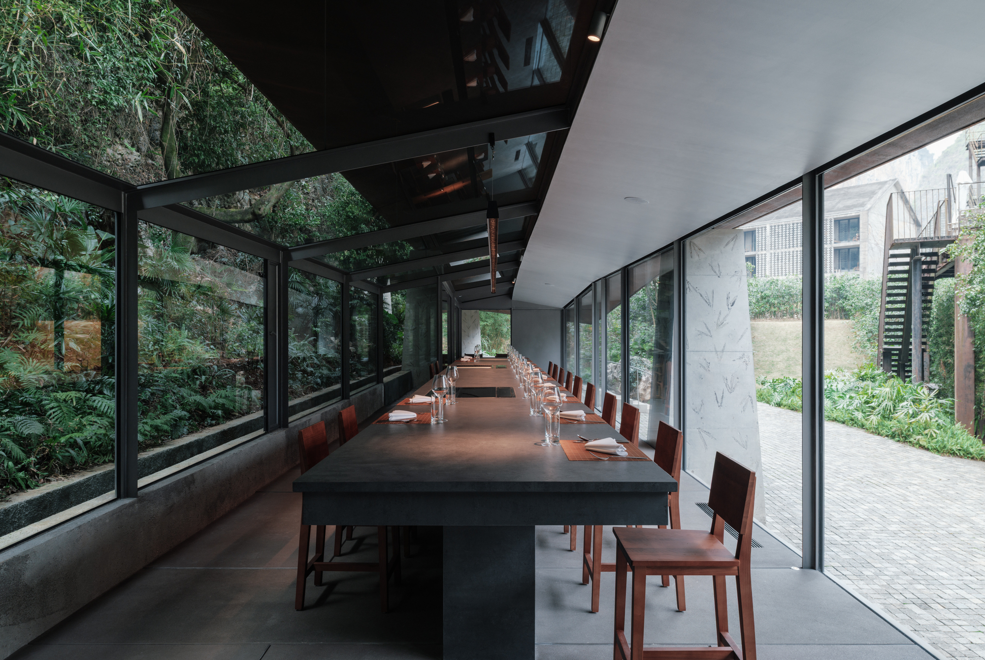 Long dining table with red wooden chairs inside a glazed pavilion, with fern-covered rock on one side and a paved courtyard on the other