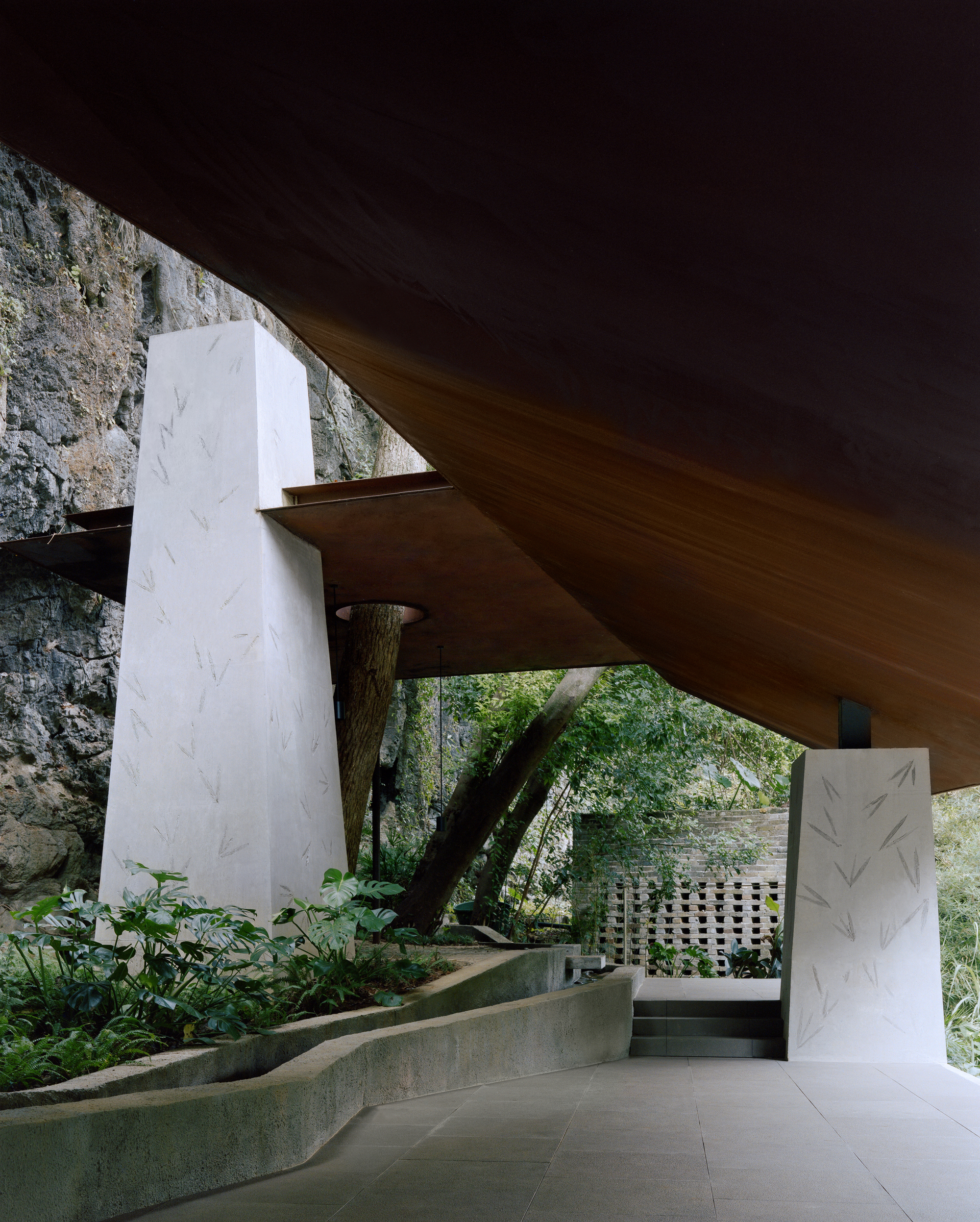 Tapered white concrete piers with leaf imprints under the sloping timber soffit, a tree trunk and perforated brick screen beyond
