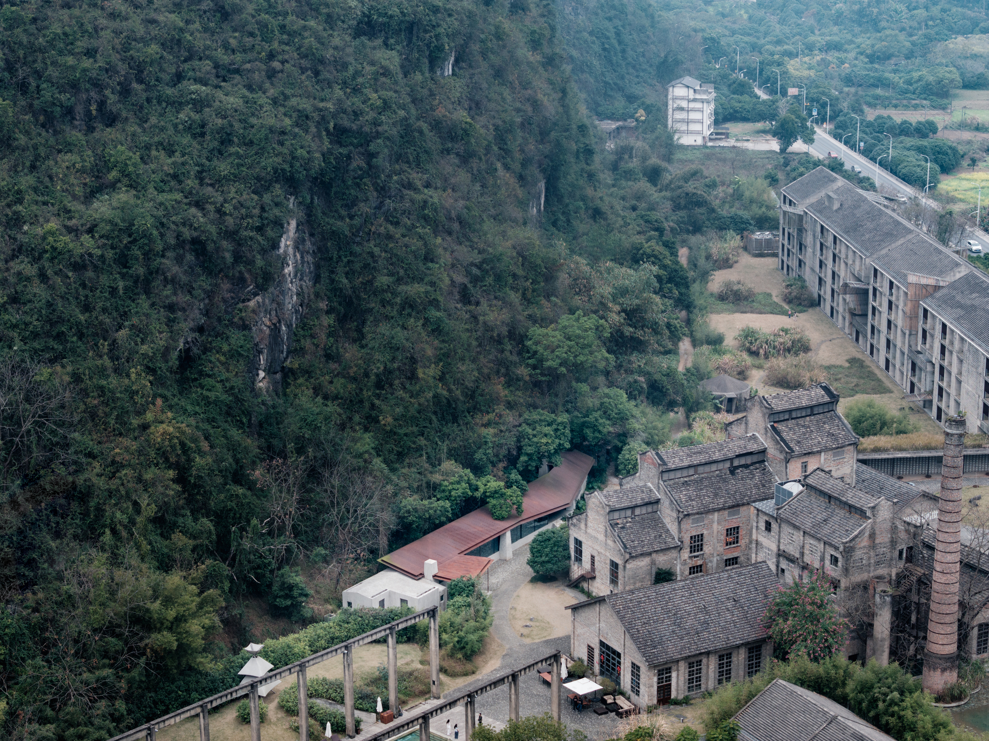 Aerial view of a rust-red sloping roof tucked between a forested cliff and old brick factory halls with a tall chimney