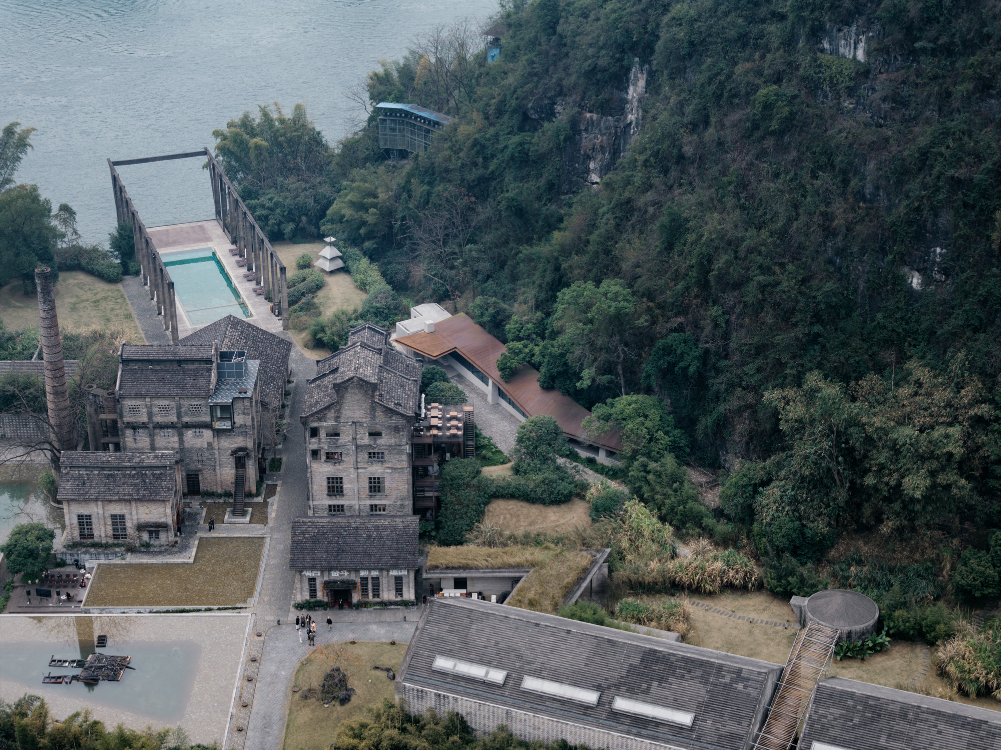 Drone view of a restored brick factory complex, a pool with timber frame and a rust-colored roof beside a river