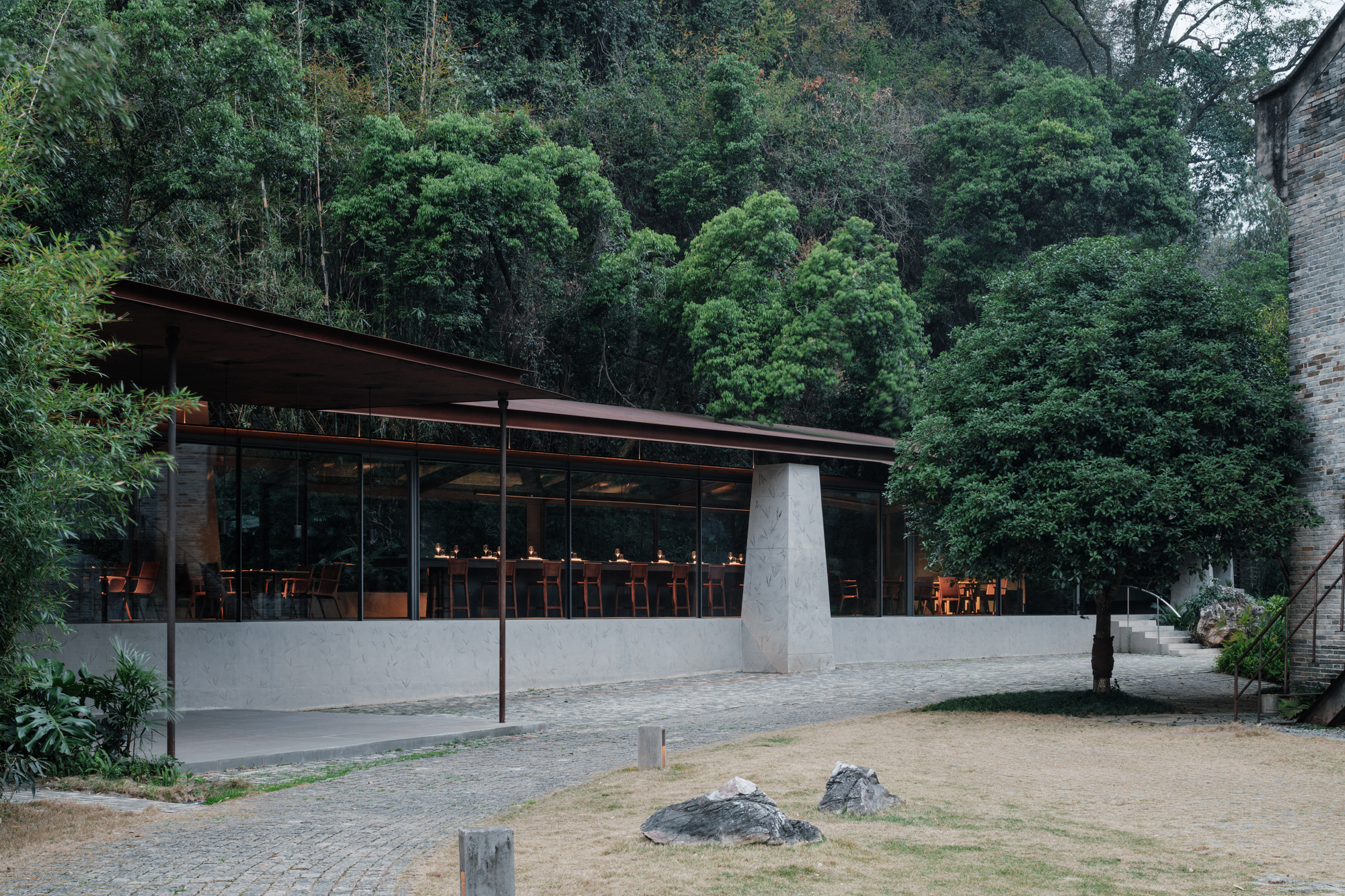 Glazed restaurant pavilion with a low steel roof and tapered concrete pier, diners lit by warm lamps against dense green hillside