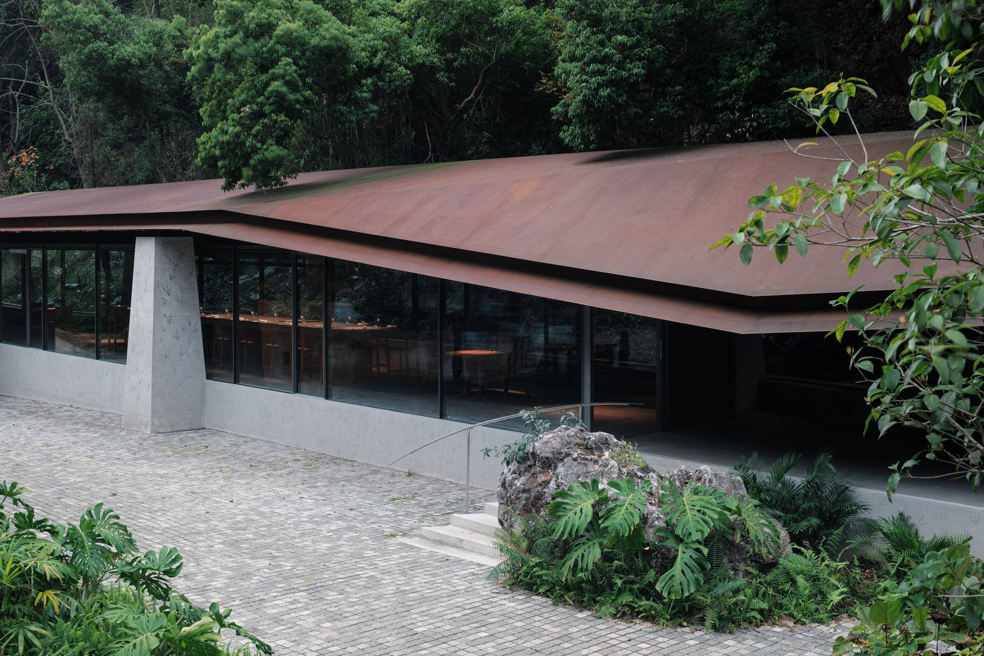Weathering steel roof folding over a glass-walled pavilion, with concrete pier, stone steps and a planted rock bed in the foreground