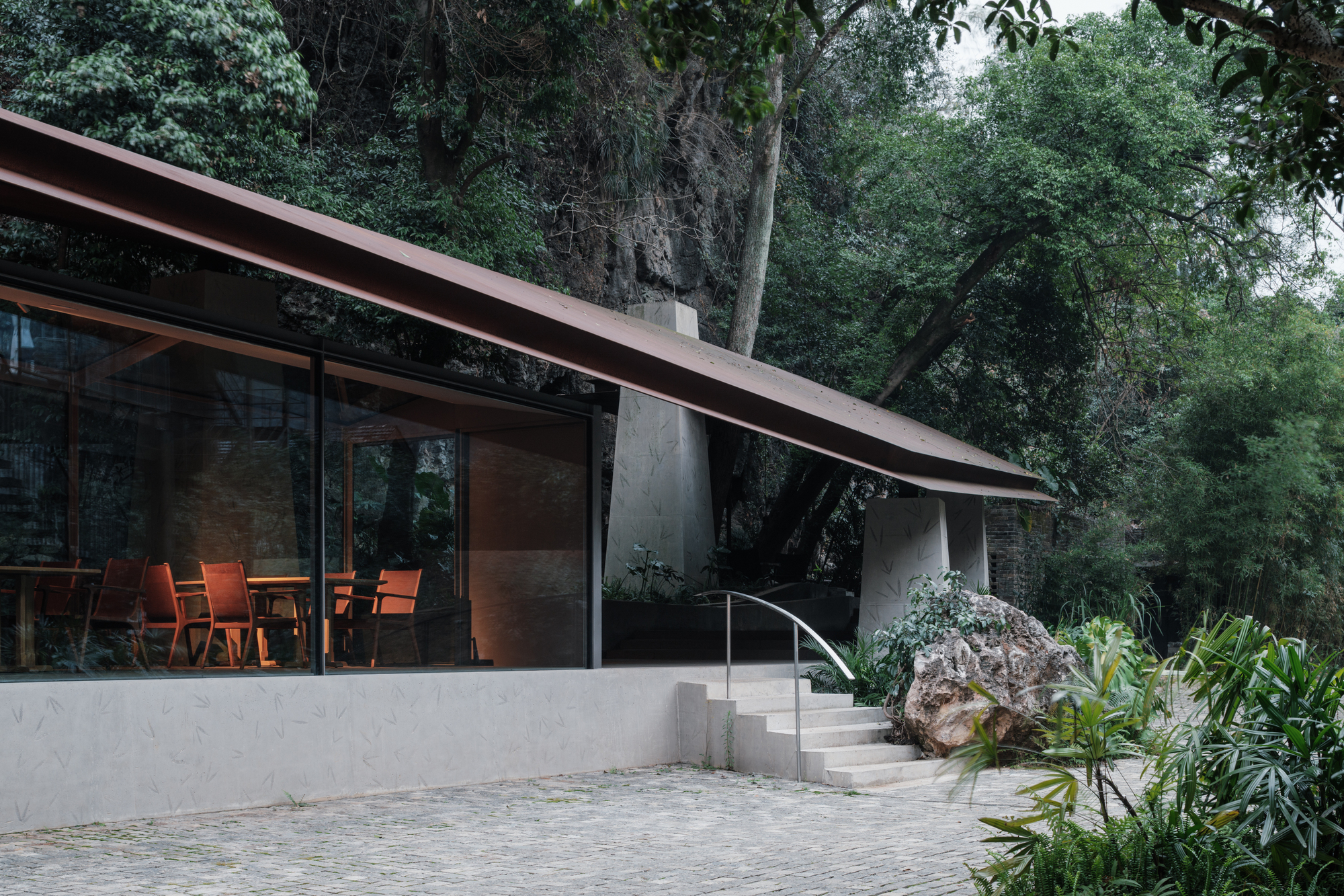 Side view of the glazed dining room under a thin tilted steel roof, with orange chairs visible and steps rising beside a boulder