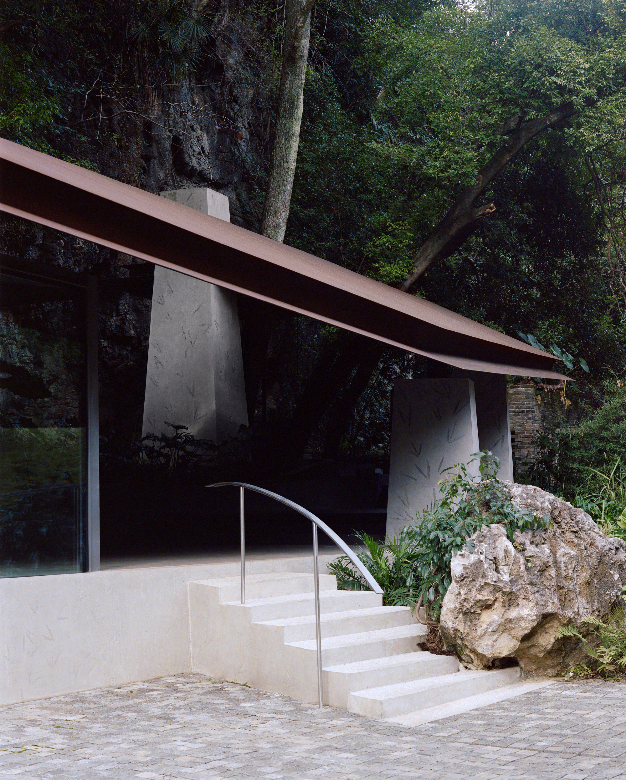 Concrete steps with a curved steel handrail rising past a limestone boulder to tapered piers under the thin rust-colored roof edge